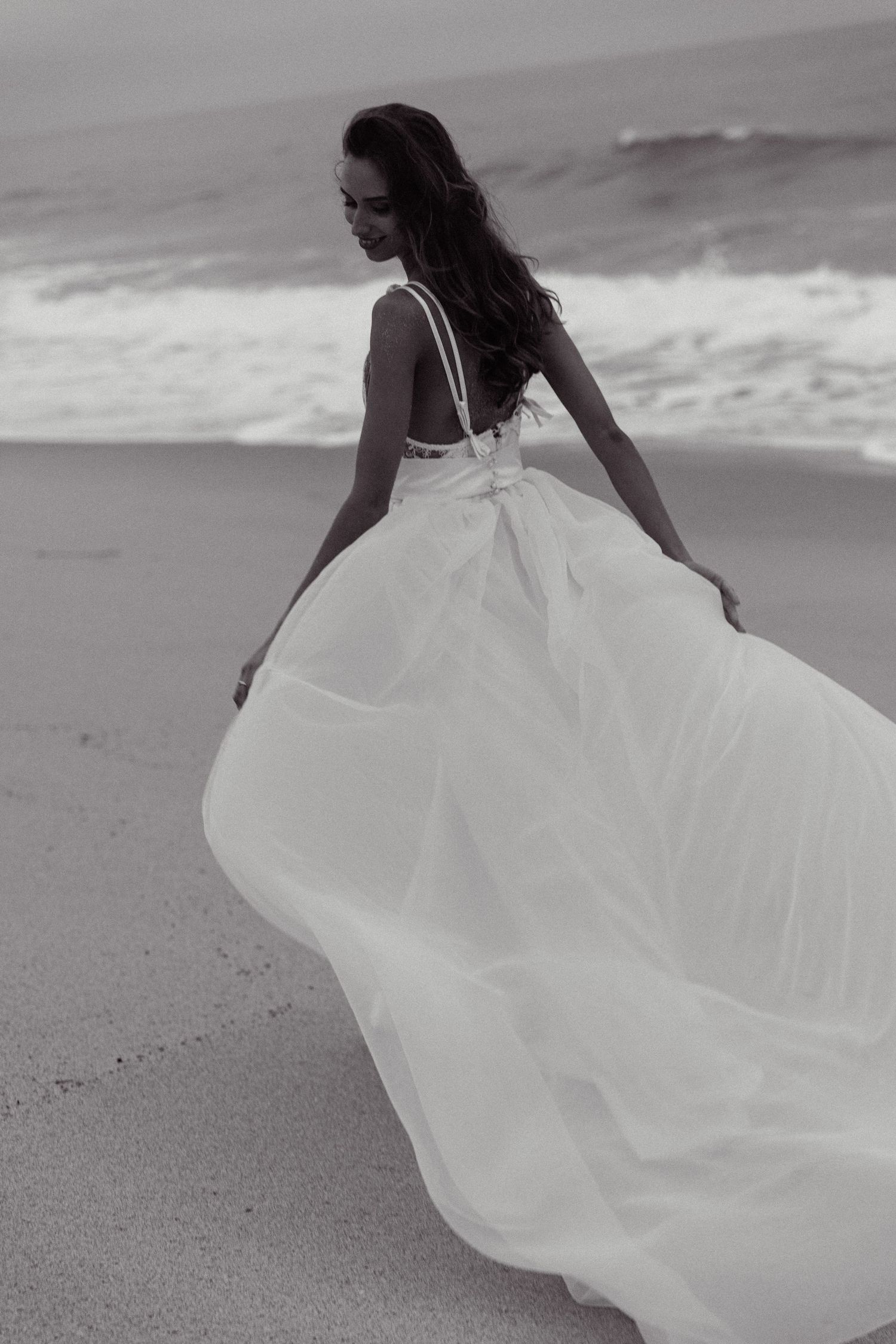 Dramatic black and white photo of bride in flowing dress twirling on beach with ocean waves.
