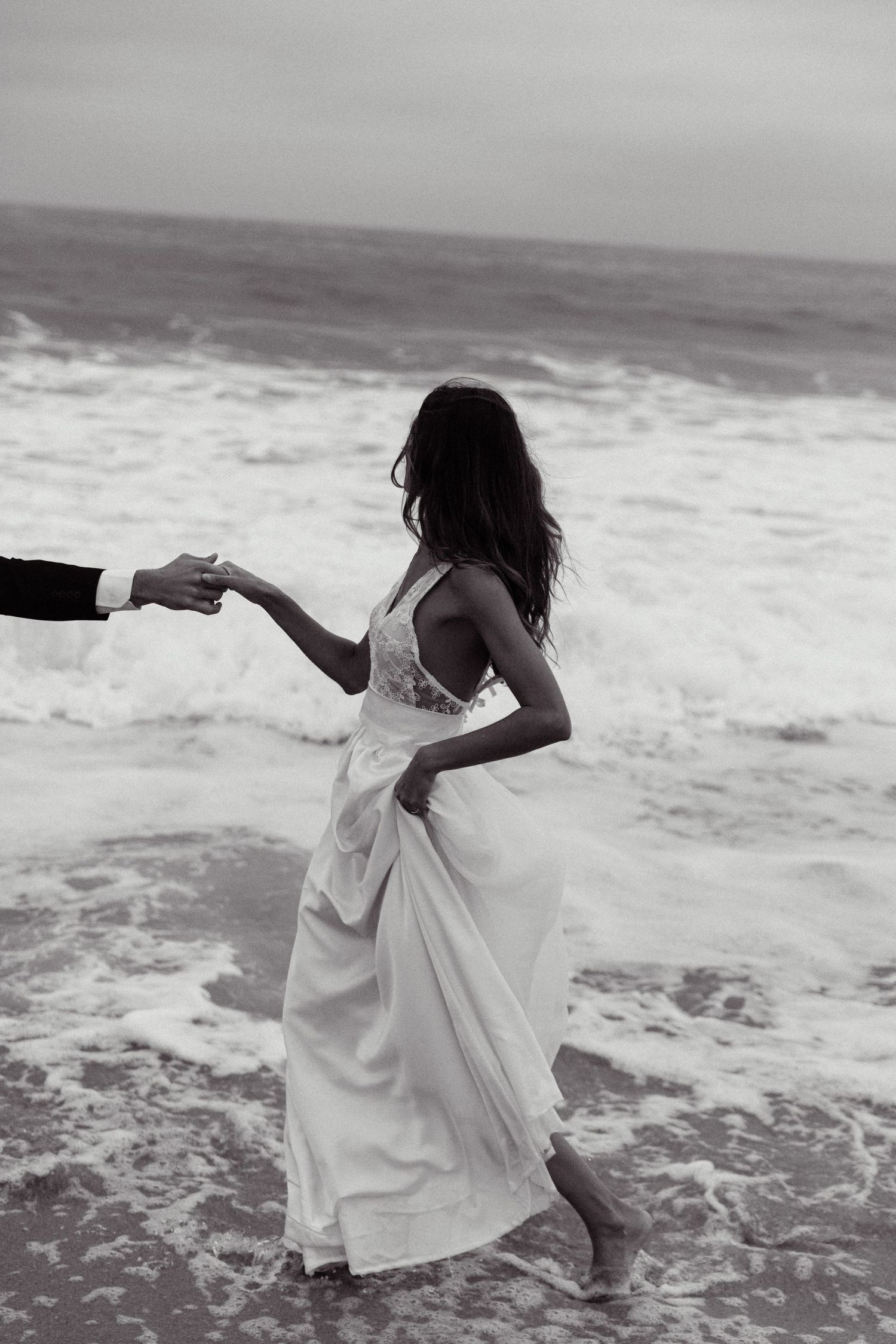 A woman in a flowing white dress dances on a beach while holding hands with someone out of frame by the ocean waves.