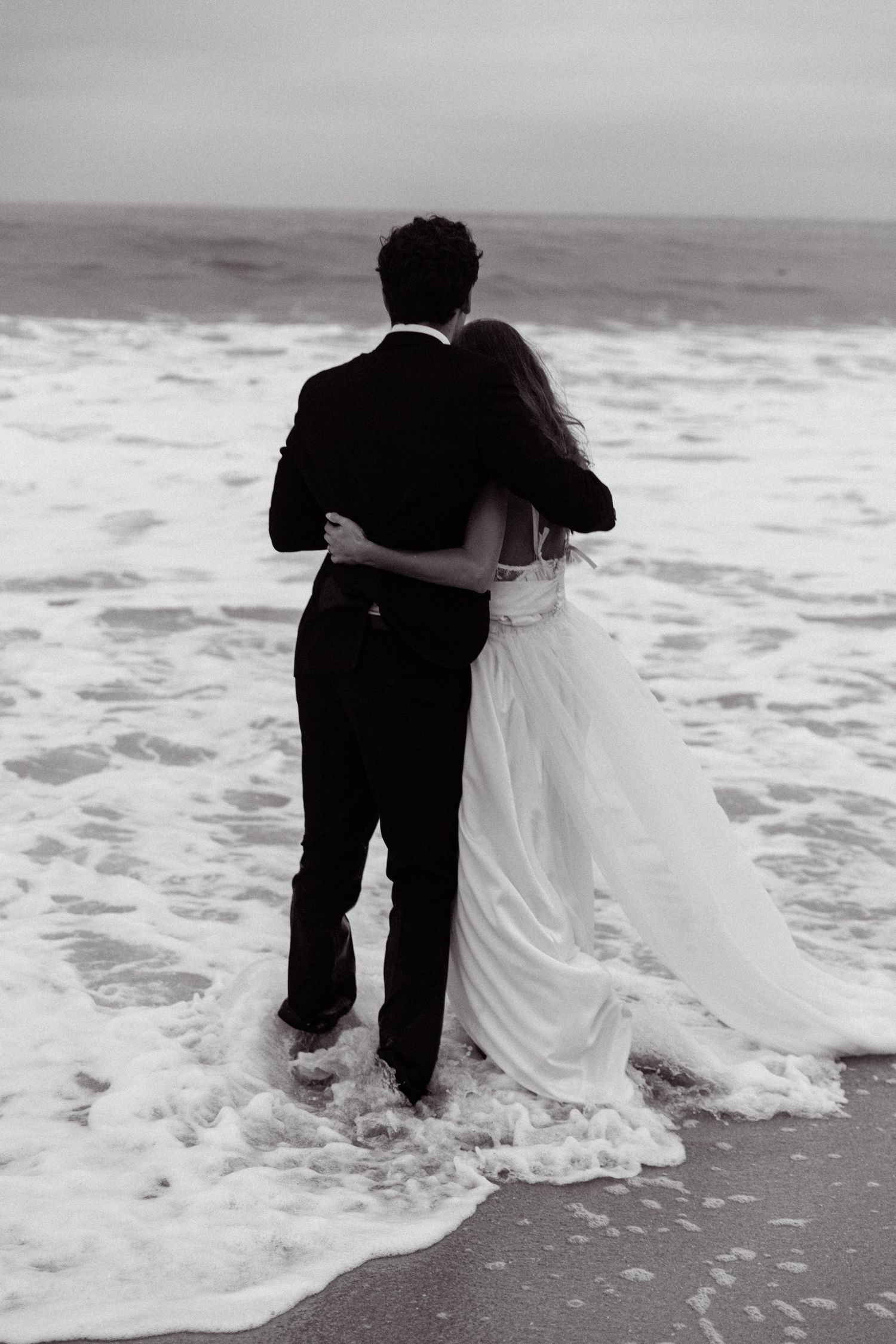 Black and white photos of newlyweds standing together in ocean waves at the beach.