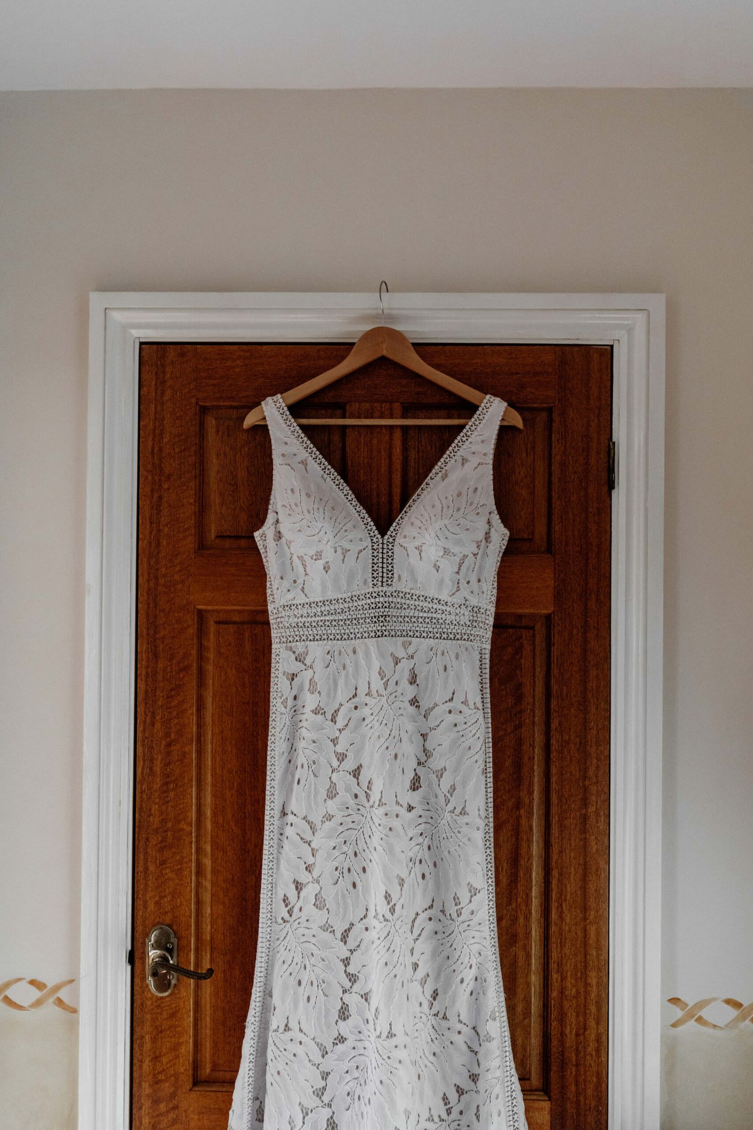Elegant white lace wedding dress hanging on wooden door against neutral wall.
