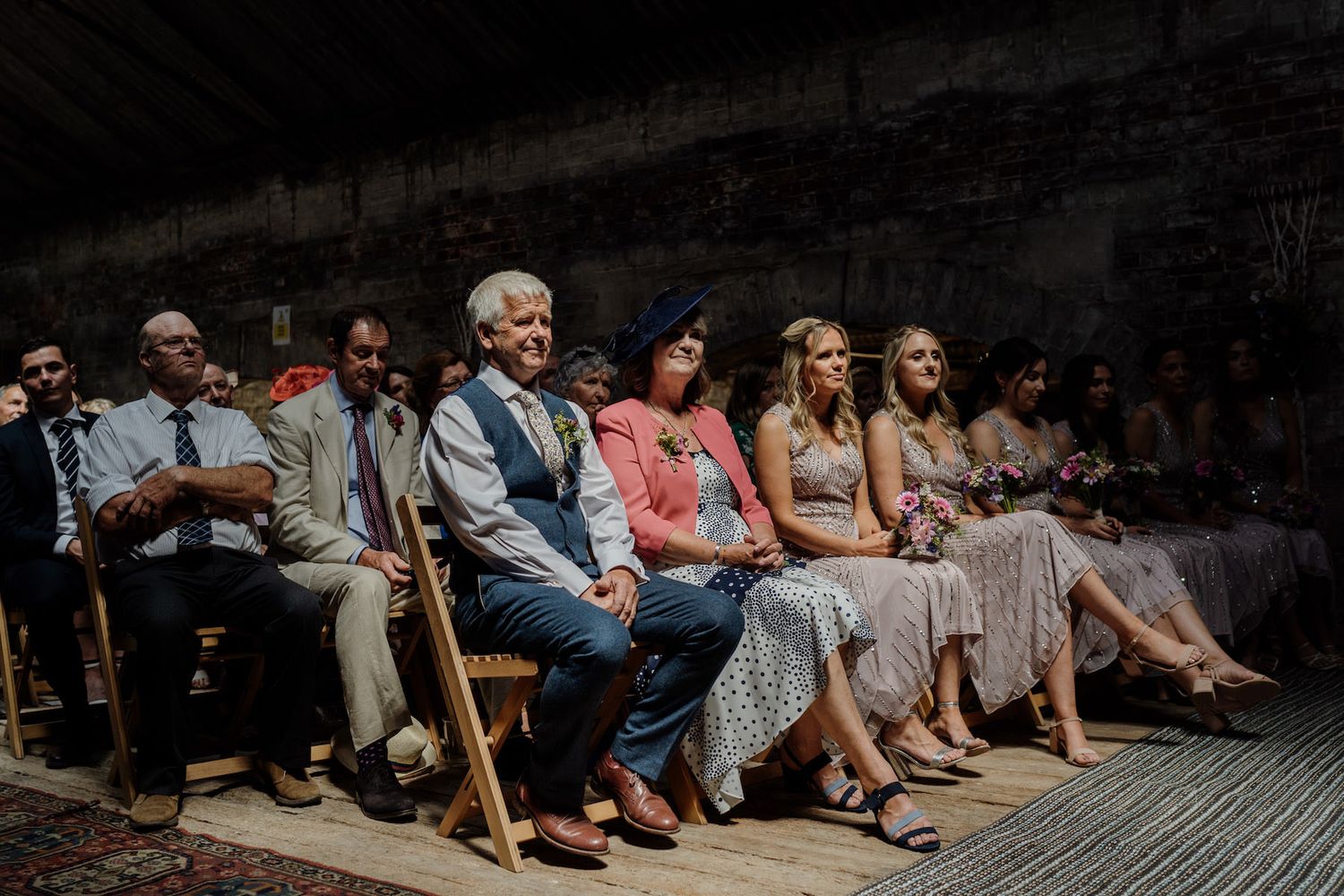 Wedding guests seated on wooden chairs in a rustic indoor venue.