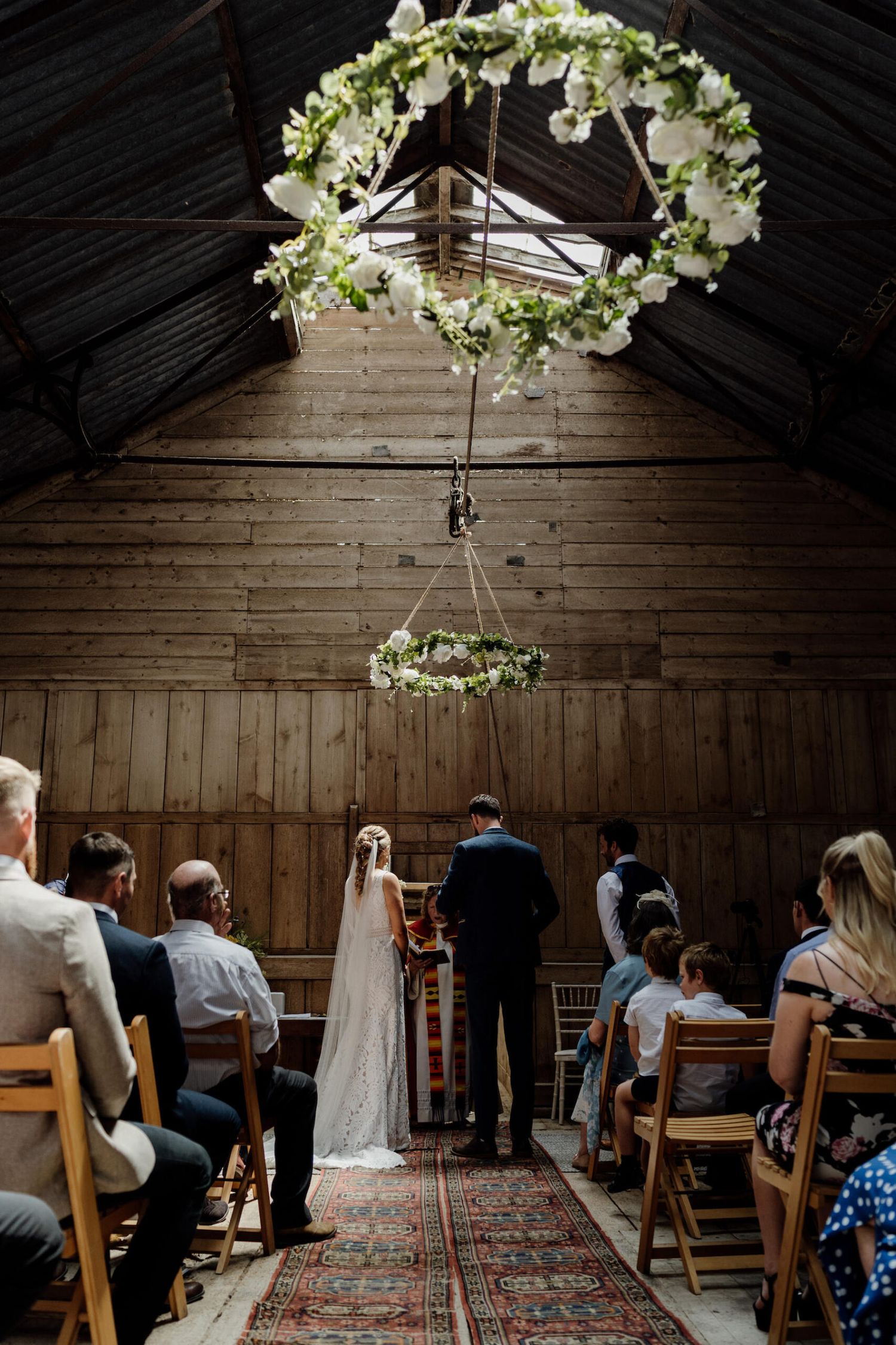 Wedding ceremony in a wooden barn under a floral arch with guests seated on wooden chairs.