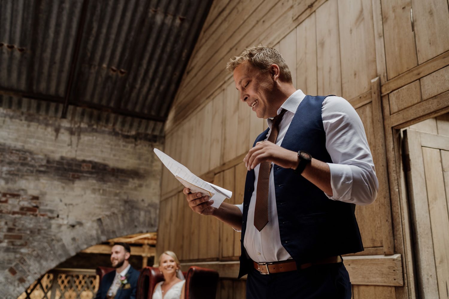 A person in a vest and tie reads from papers during what appears to be a wedding speech.