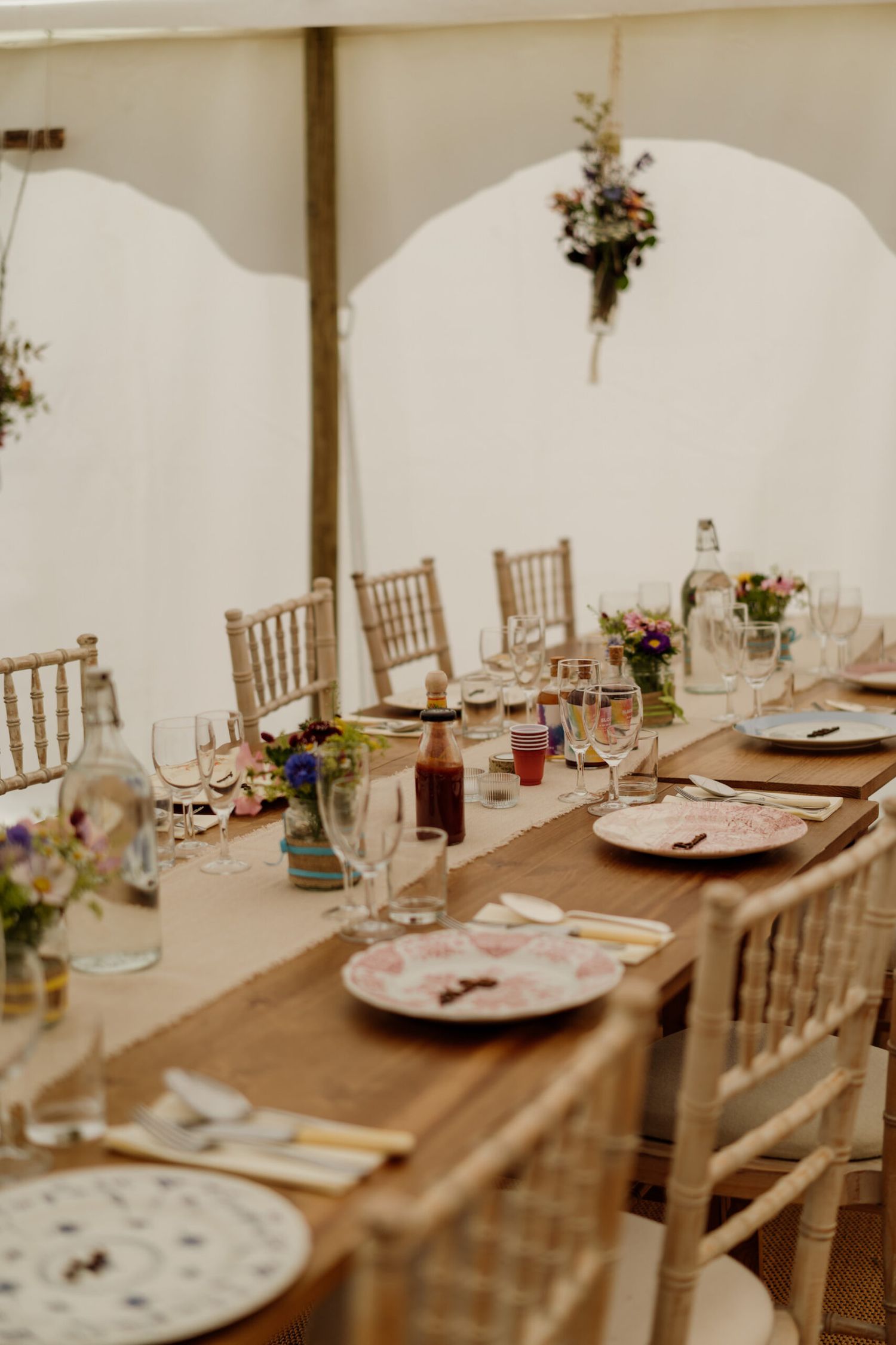 Long wooden tables set for wedding reception with vintage plates and floral centerpieces in a white tent.