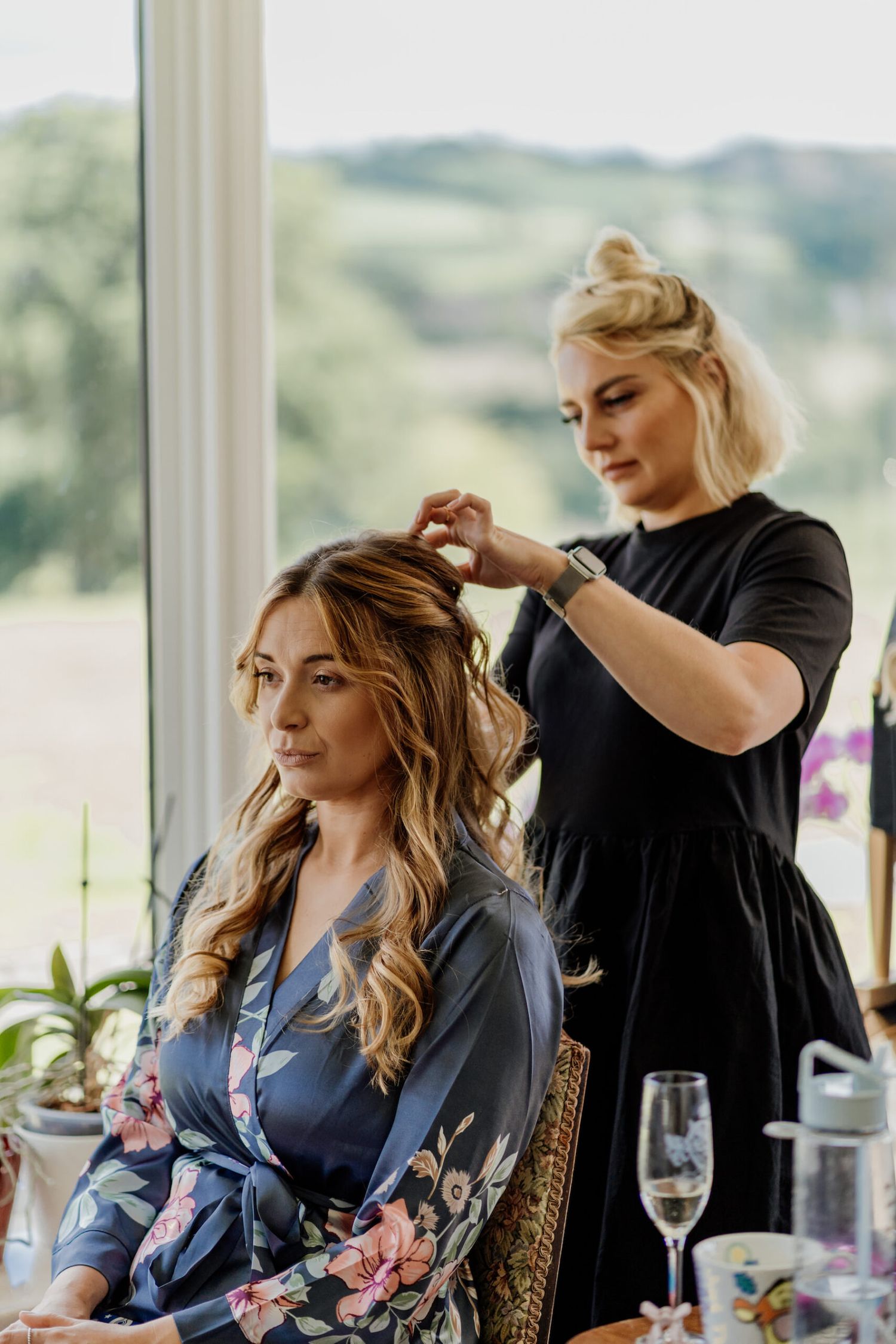 A hairstylist works on styling someone's long blonde hair in a sunlit room with large windows.