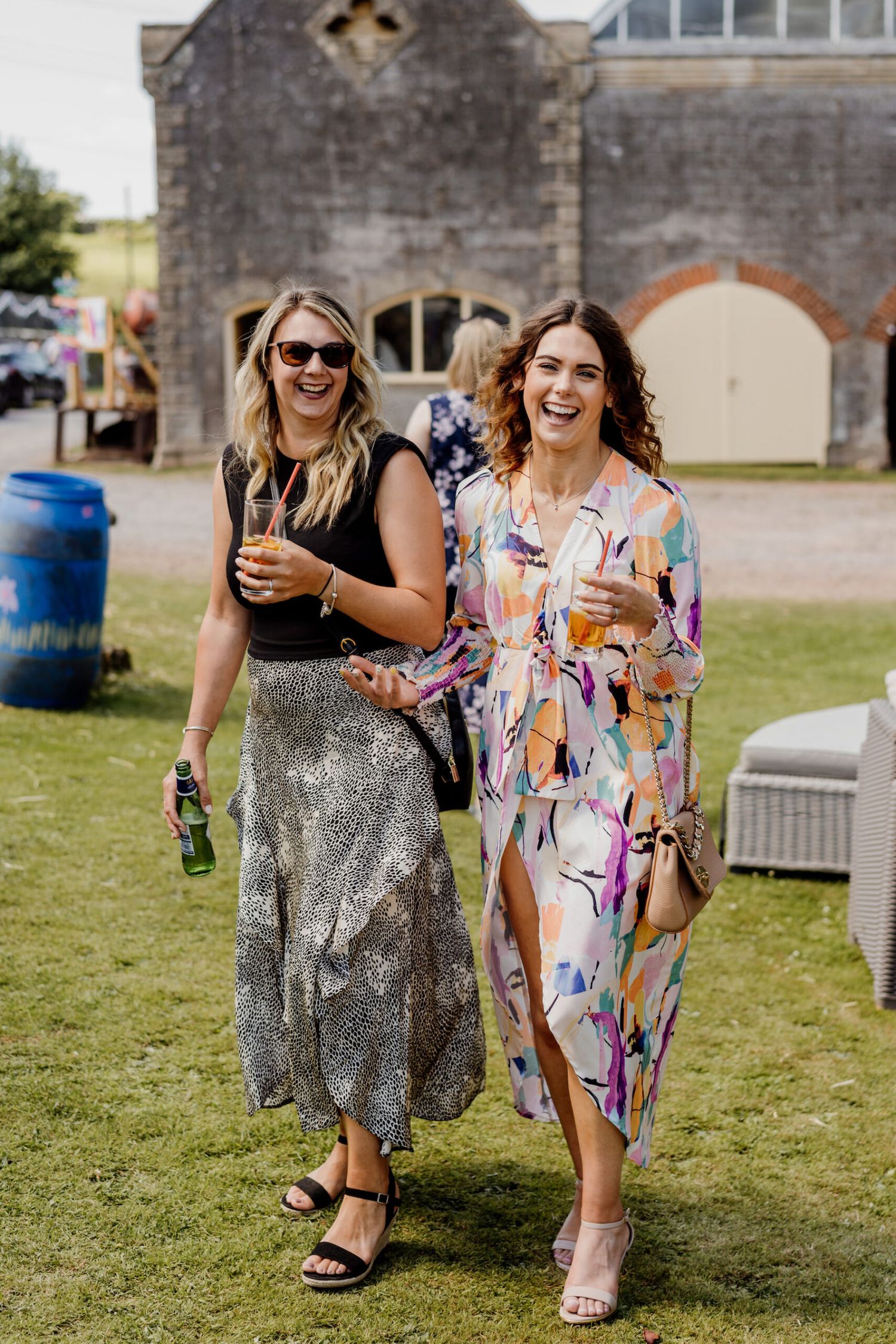 Party guests in sparkly and floral dresses enjoying drinks on a sunny lawn.