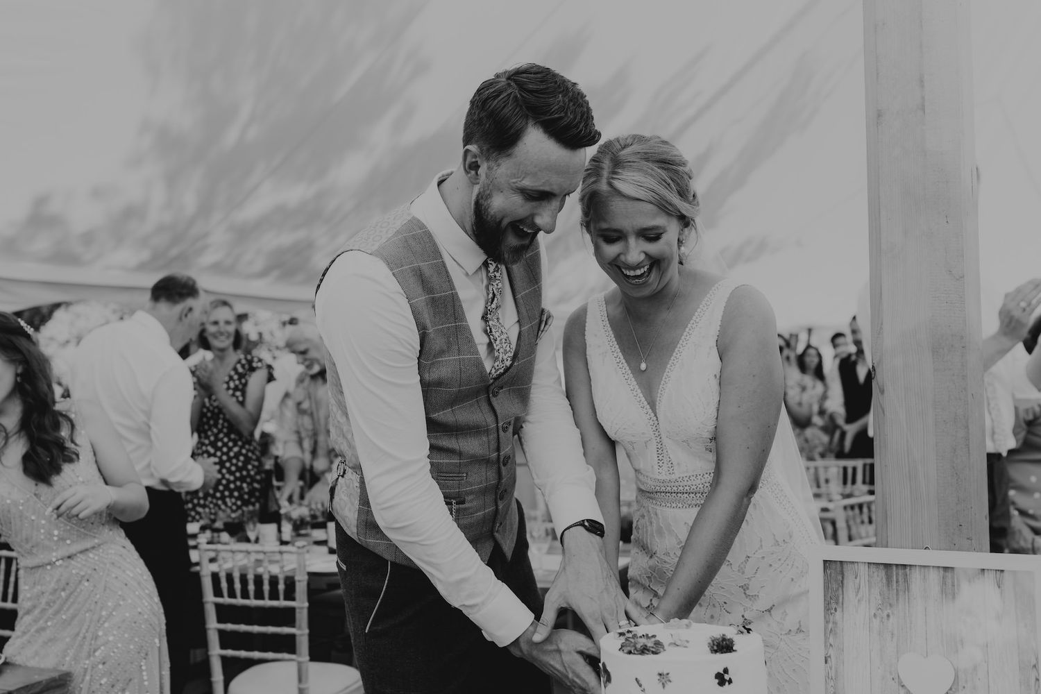 A couple in wedding attire cuts their cake together under a white tent during their reception.