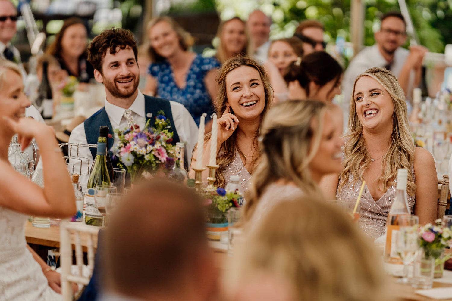 Wedding guests laugh and smile during an outdoor reception celebration with colorful flowers and wine glasses on tables.