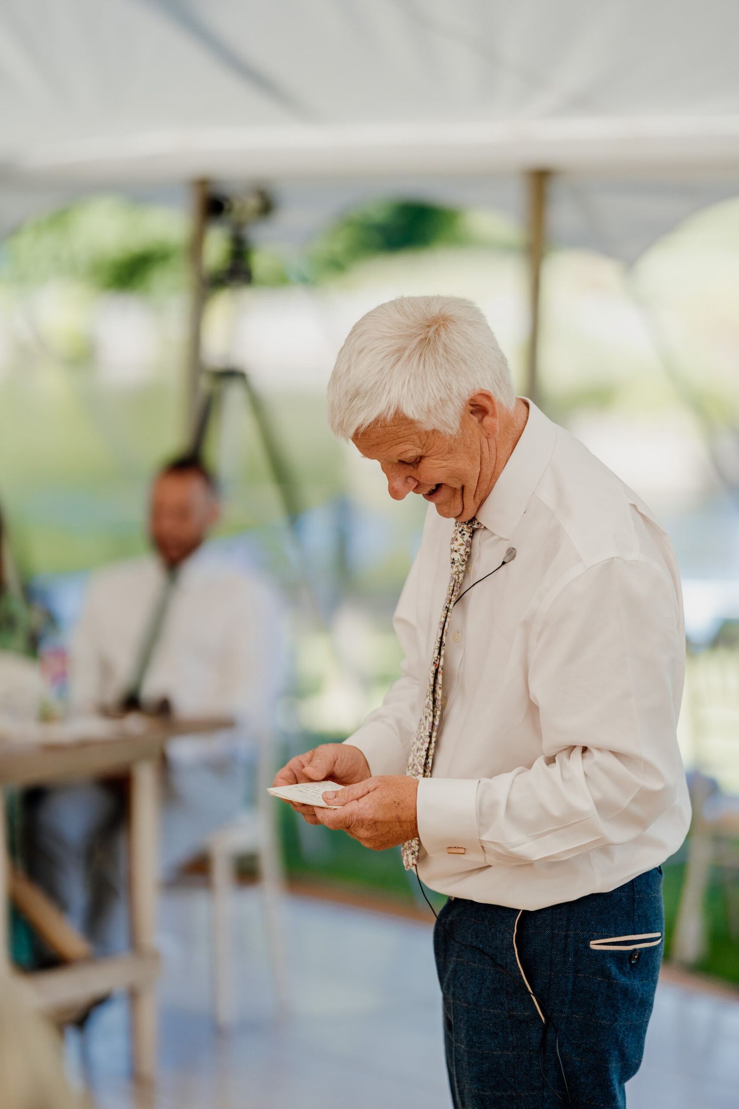 An elderly guest in a white sweater checks his phone during a wedding reception under a tent.