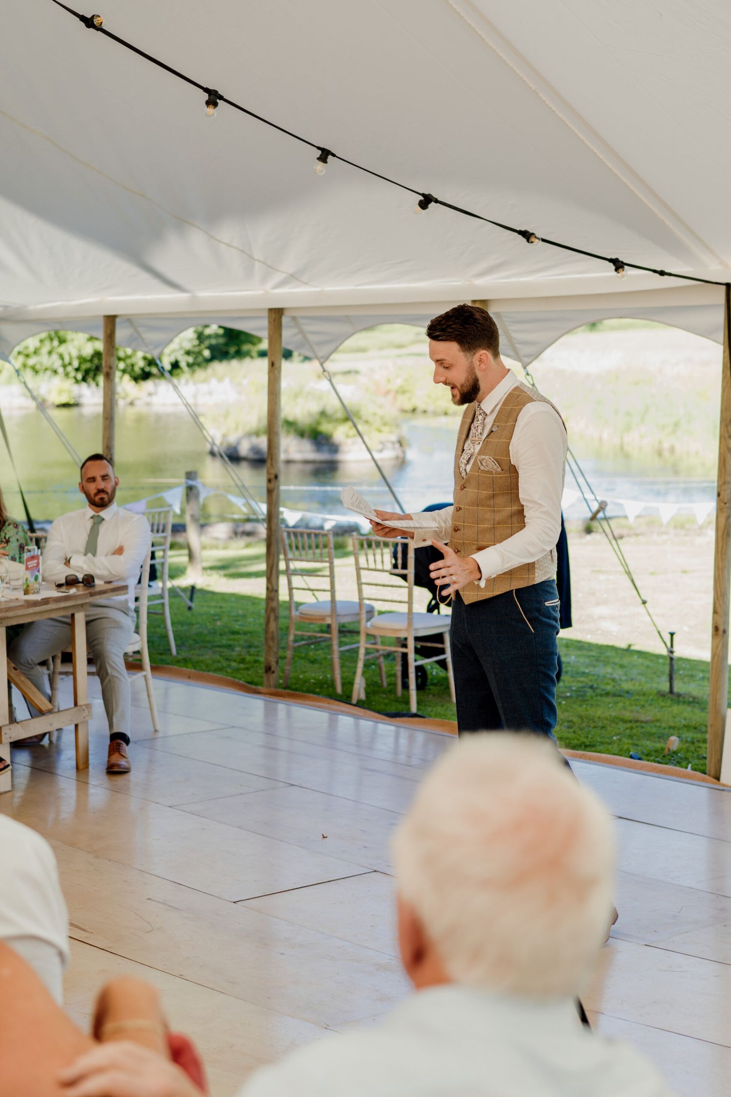 A person in a tan vest gives a speech under a tent during a wedding reception with string lights overhead.