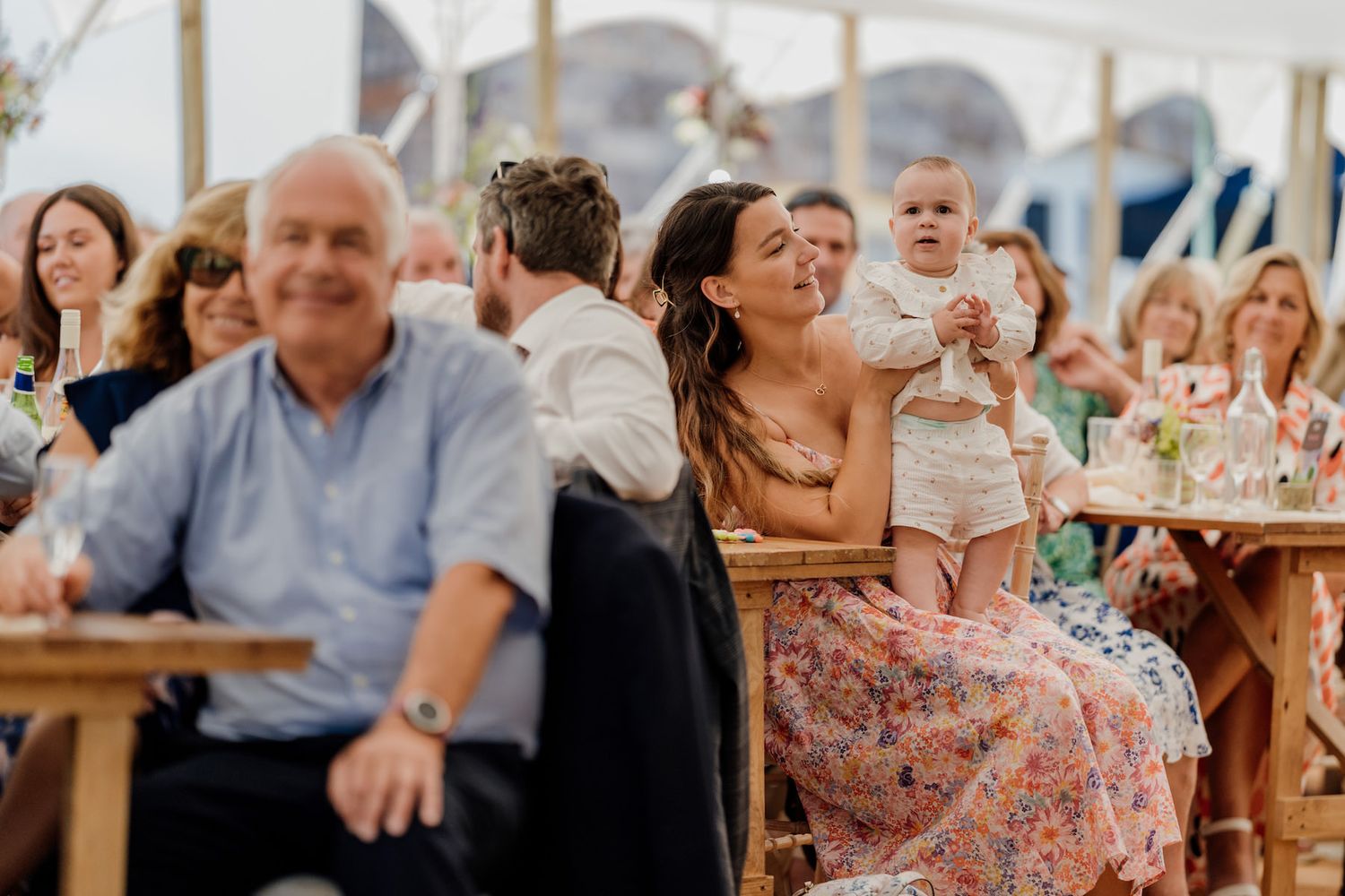 Wedding reception guests react with laughter while seated at wooden tables during celebrations.
