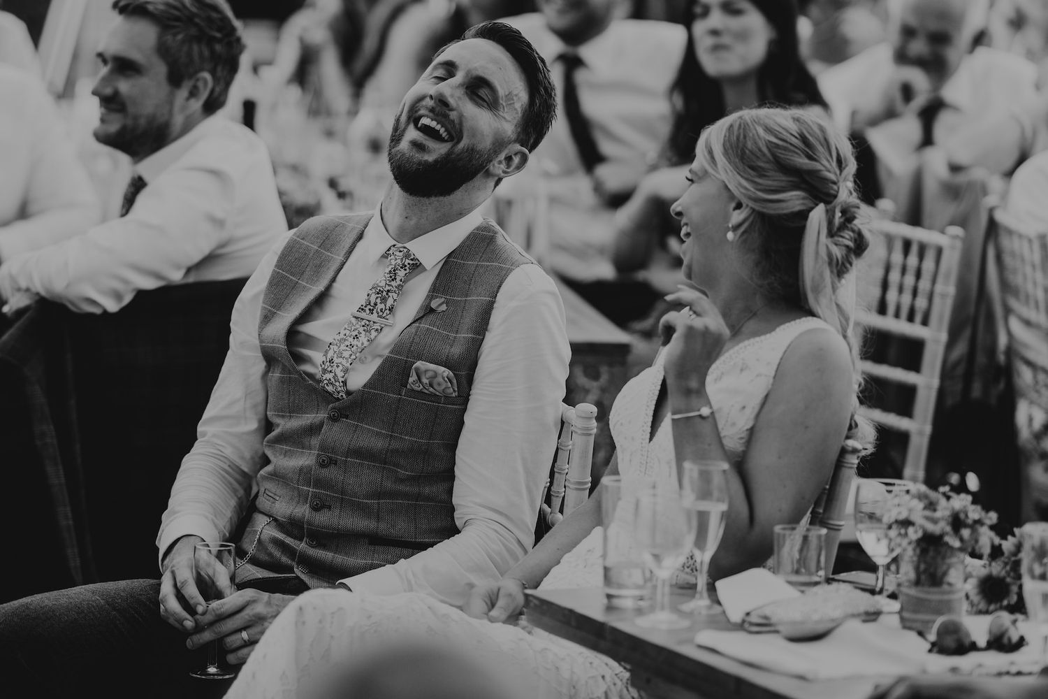 A black and white photo captures genuine laughter during wedding reception speeches.