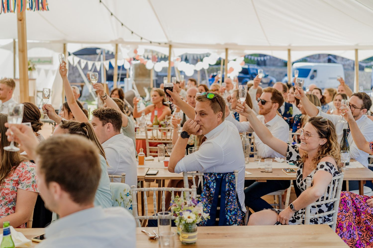 Wedding reception guests raise their hands in celebration under a white tent with string lights.