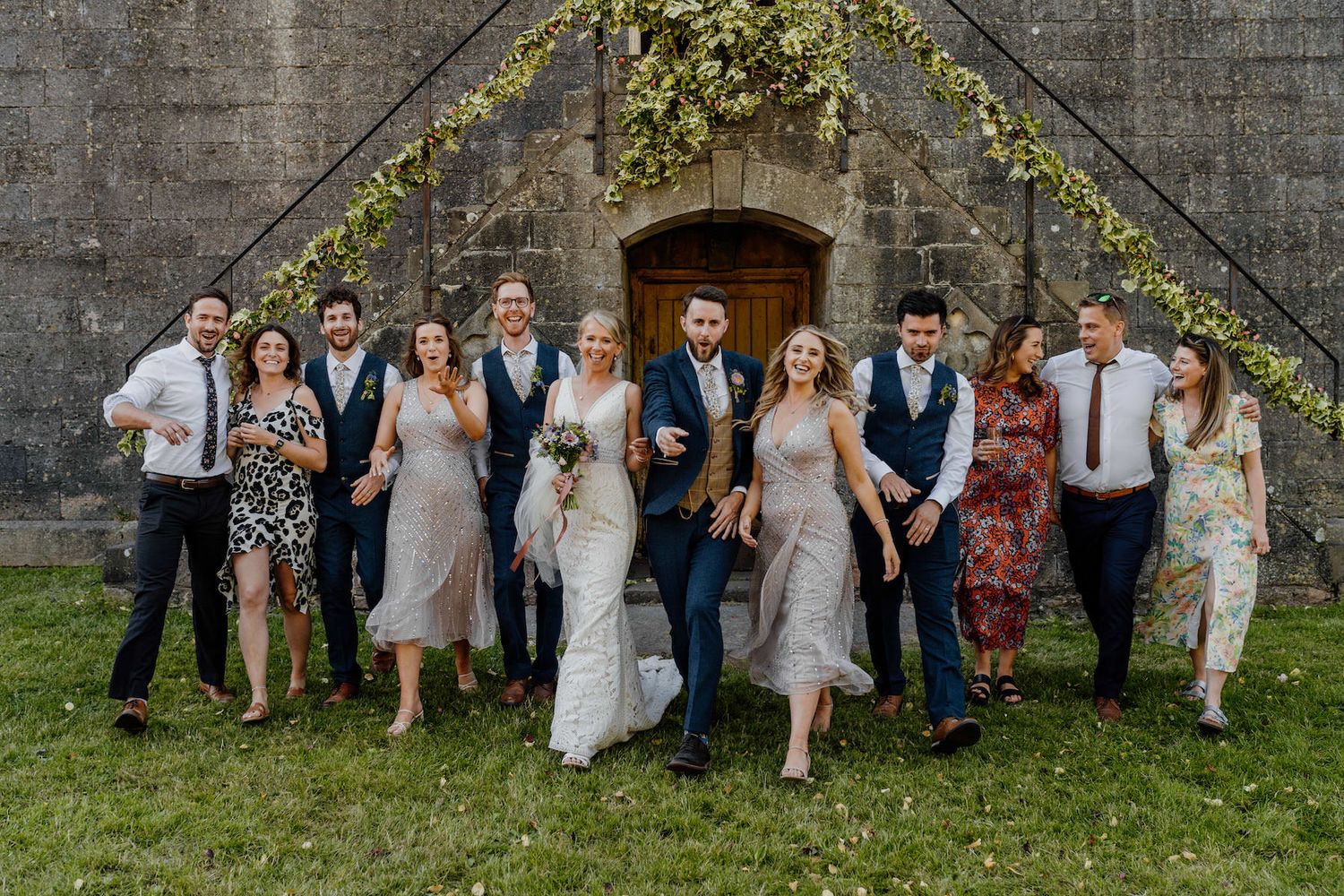 Wedding party poses together against stone wall backdrop with greenery decorations above.