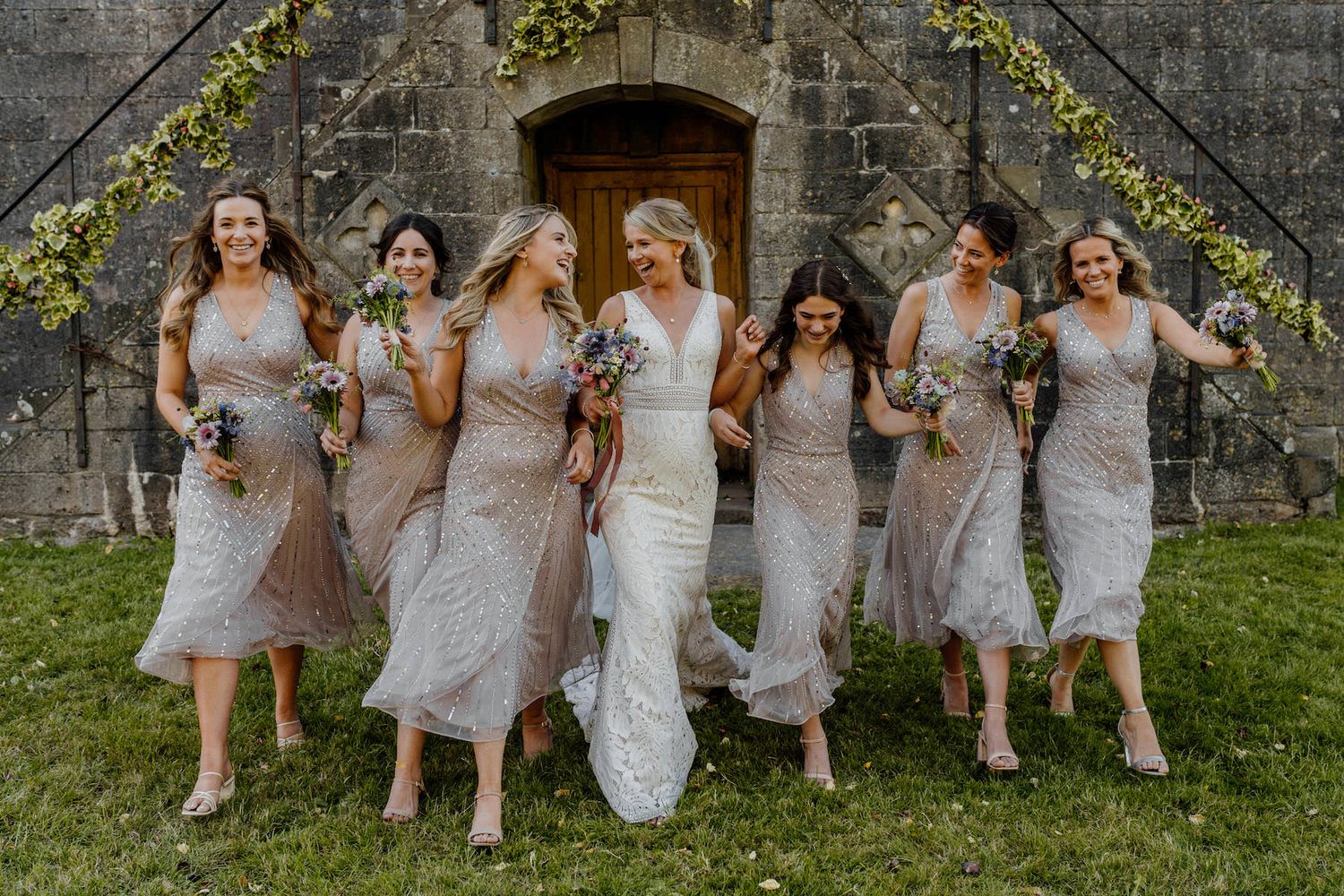 Wedding party in grey dresses and navy suits arranged casually against rustic stone wall.