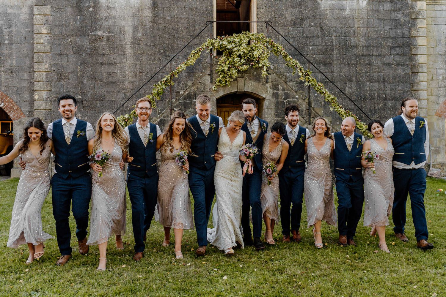 Wedding group photo with coordinated navy and grey attire against stone architecture.