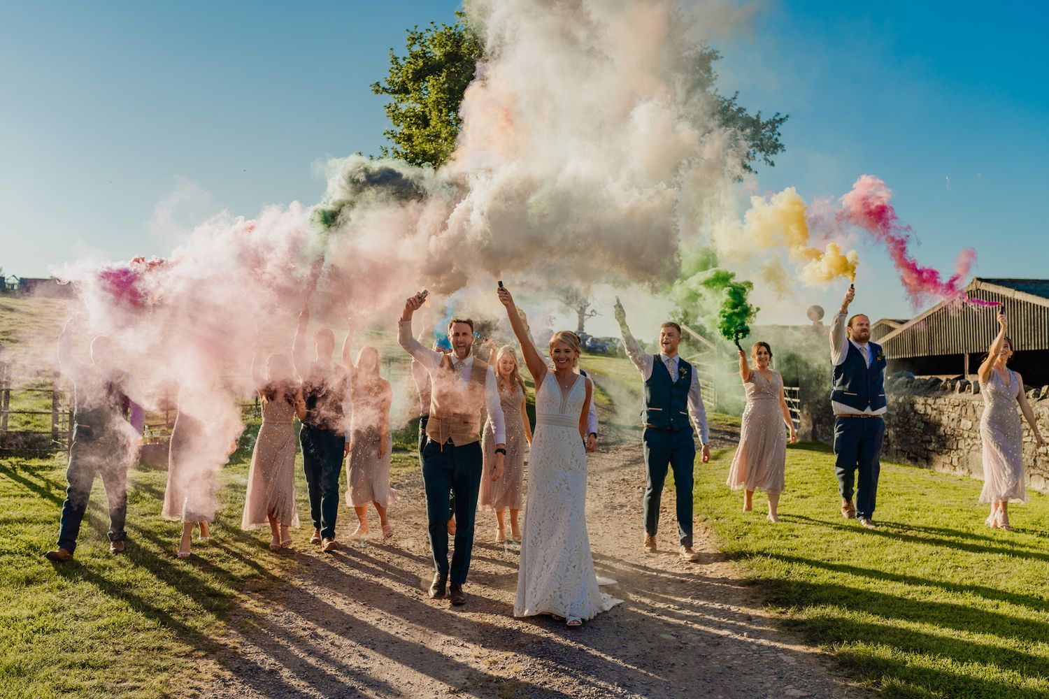 Wedding party celebrates with colorful smoke bombs on grassy field during golden hour.