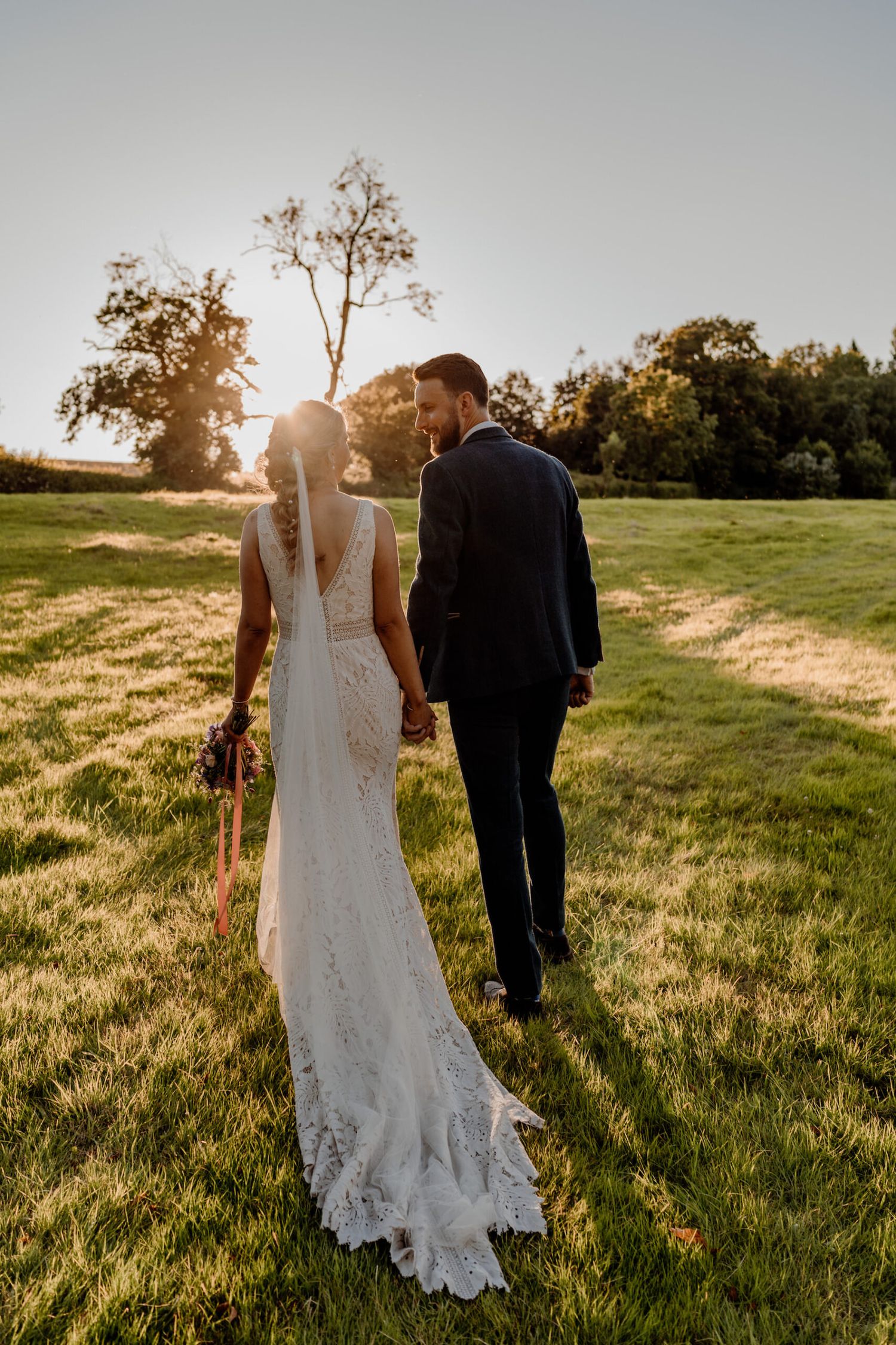 Couple holding hands while walking through a sunlit meadow in wedding attire.