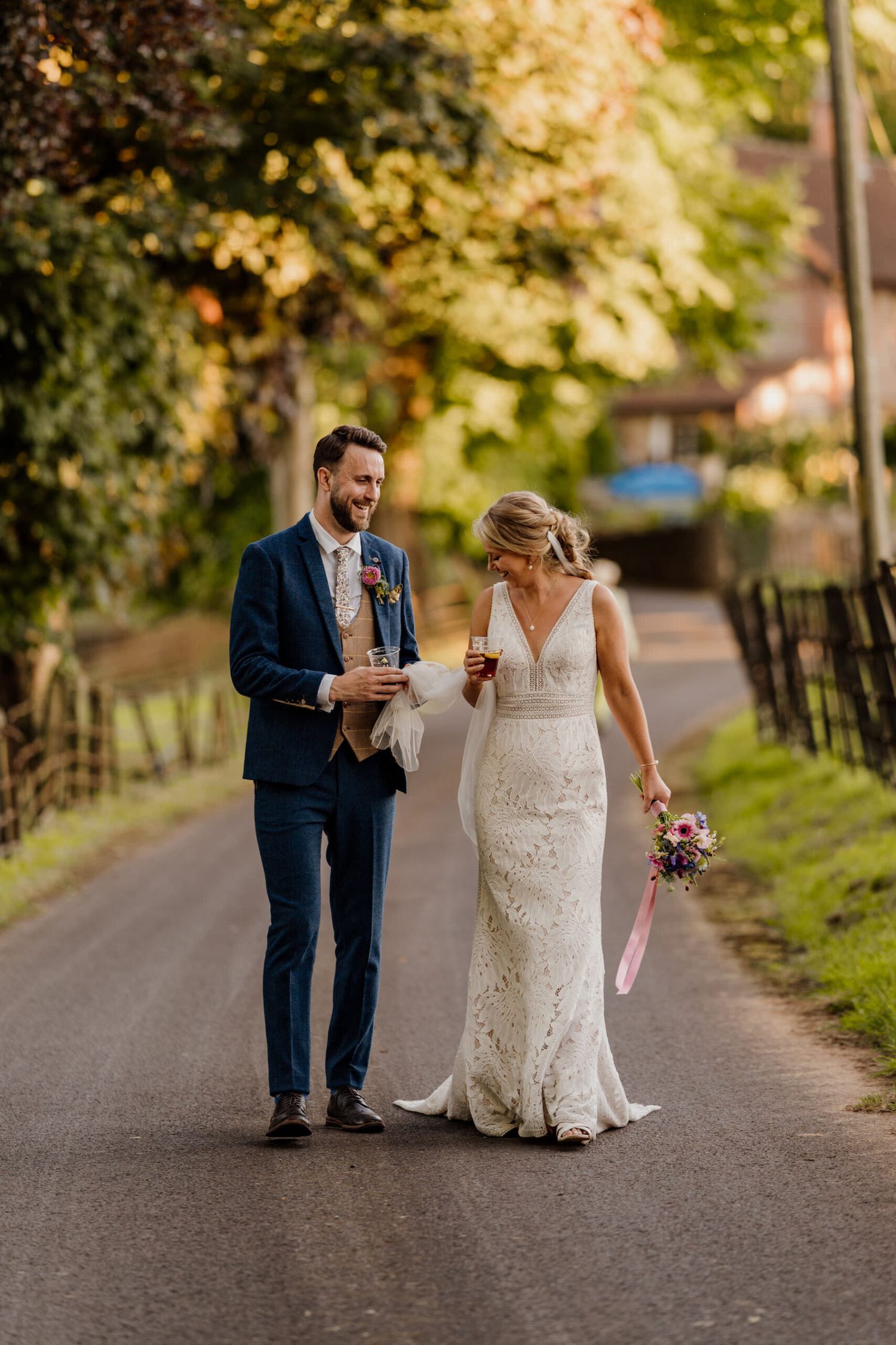 Couple walking together on tree-lined path in wedding attire during autumn.