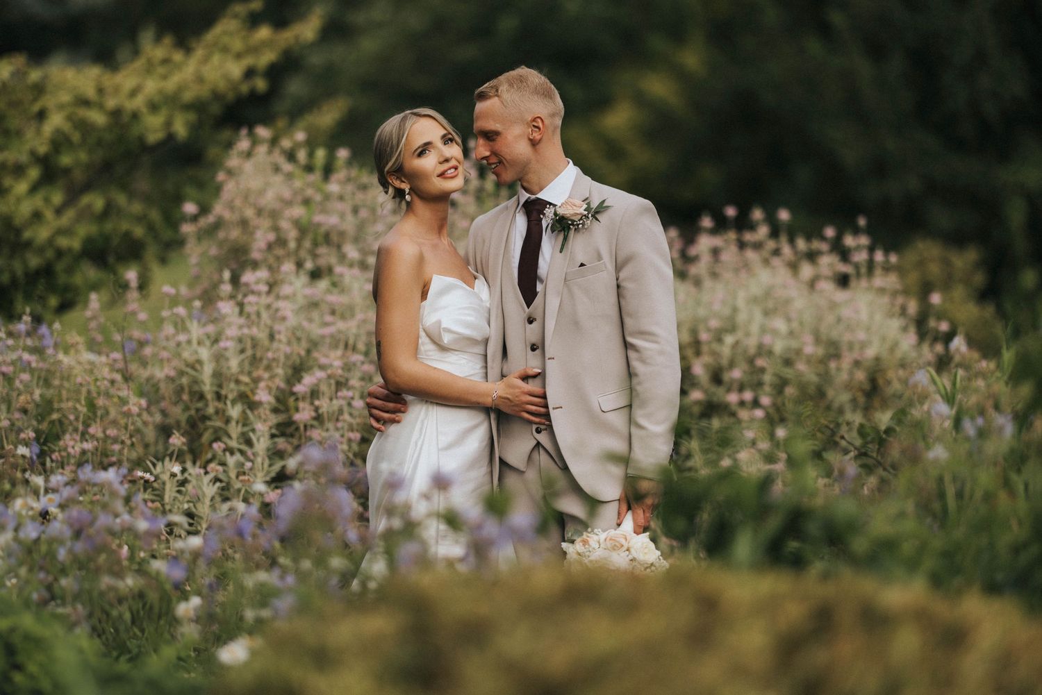A wedding couple embraces in a wildflower meadow wearing wedding attire during an outdoor photography session at Hodsock Priory.