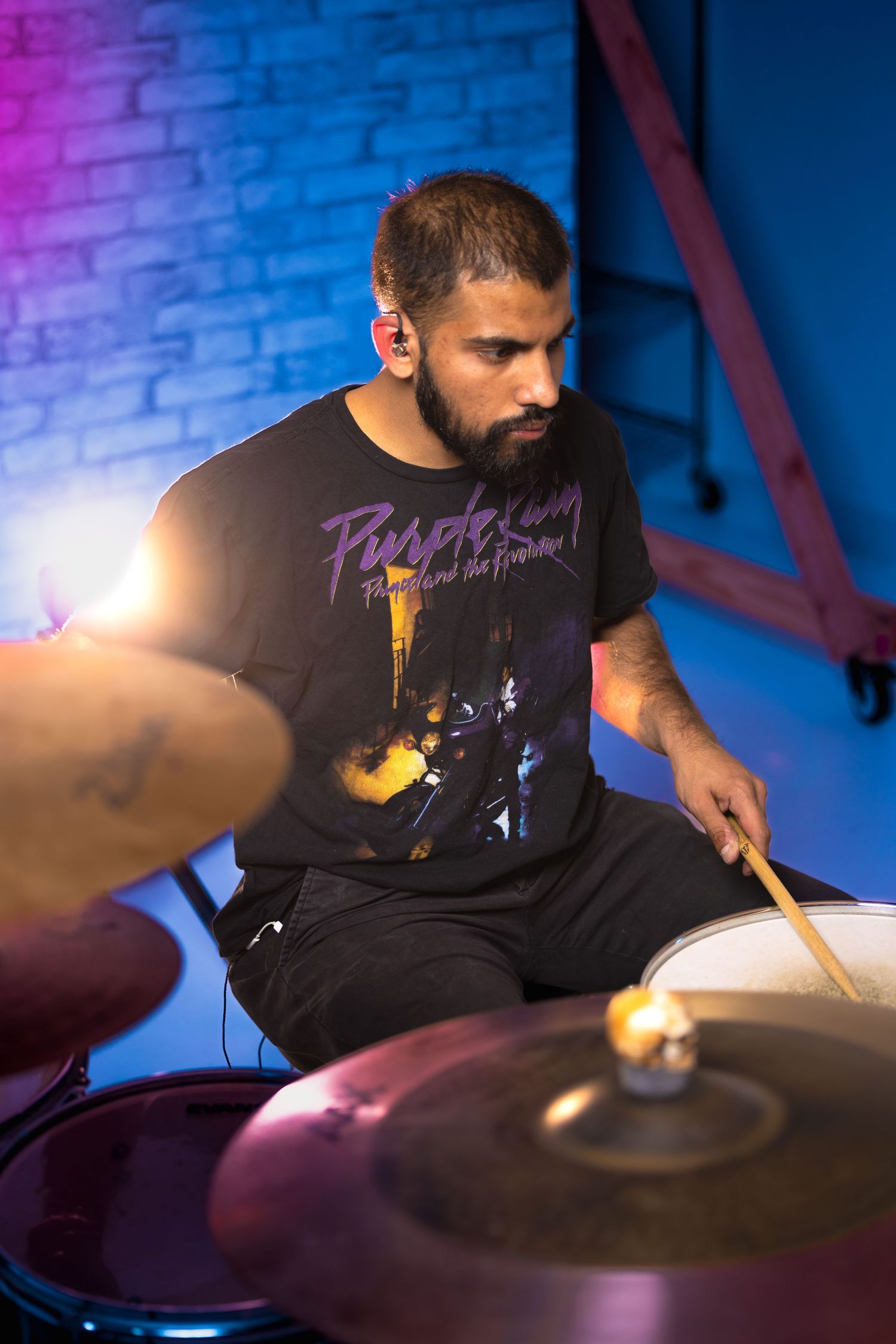 Drummer performs on stage with colorful neon lighting and brick wall background.