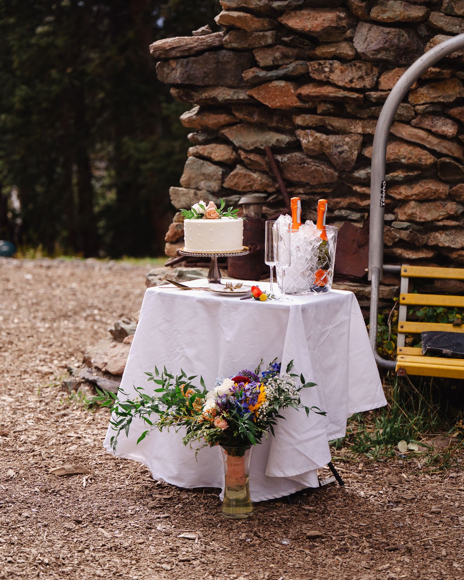 Romantic outdoor table setting with white tablecloth and floral arrangements against stone wall backdrop.