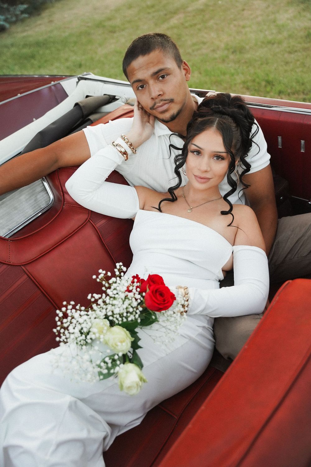 A newlywed couple poses together in a vintage red convertible car with white baby's breath flowers and red roses.