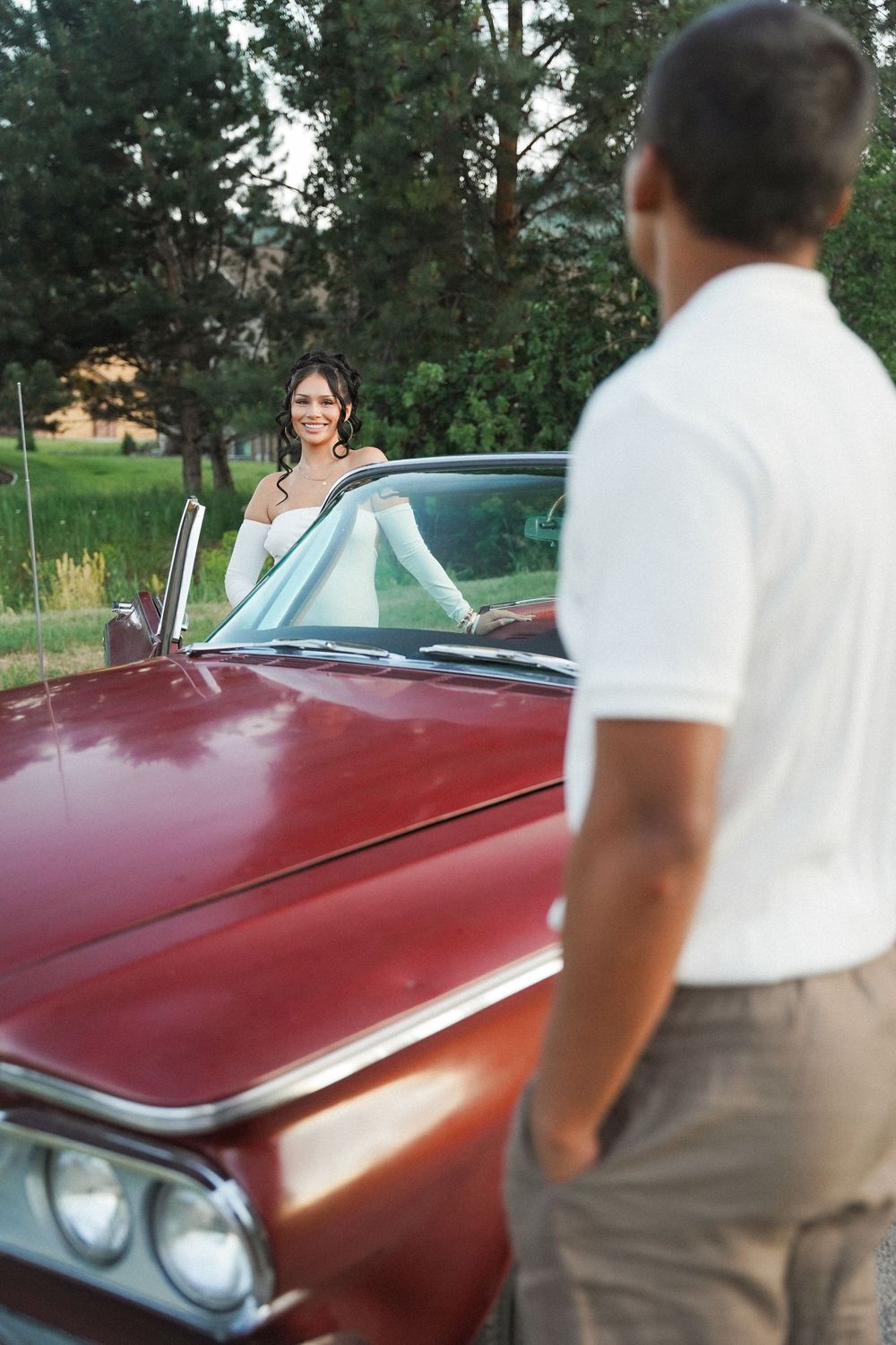 A couple poses with a classic red vintage car parked near pine trees on a summer evening.