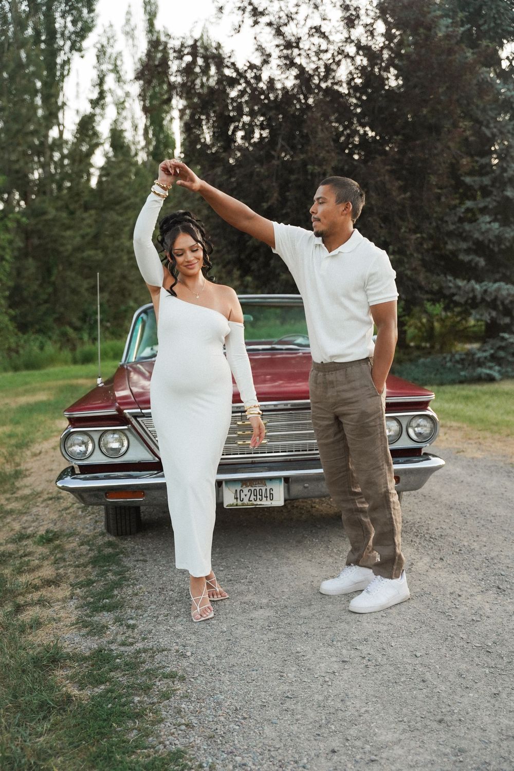 A couple in elegant white outfits dance together in front of a classic red car during a romantic outdoor photoshoot.