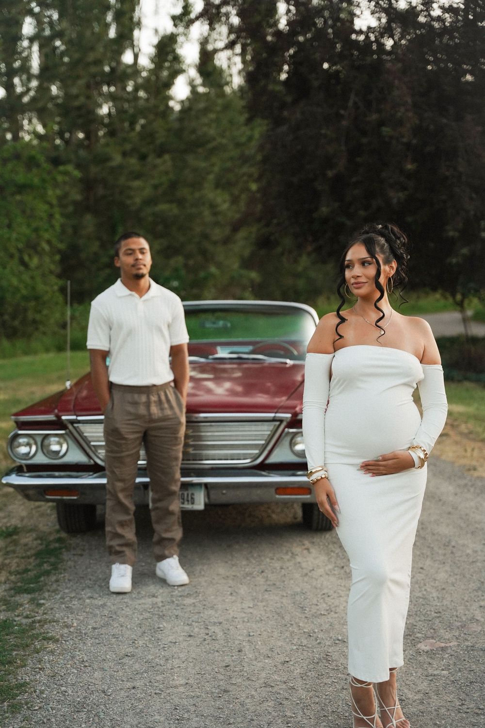 A couple poses for a vintage-inspired photoshoot in elegant attire next to a classic red car on a gravel road.