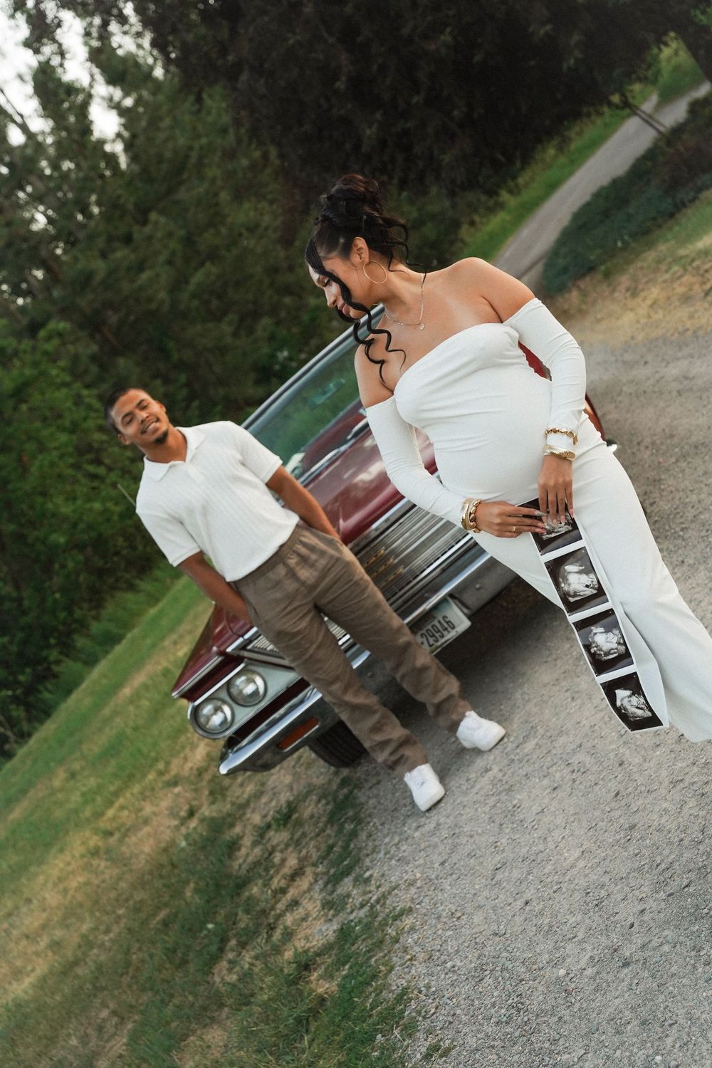 Two people pose playfully on a gravel path next to a car, with one wearing all white and the other in casual attire.