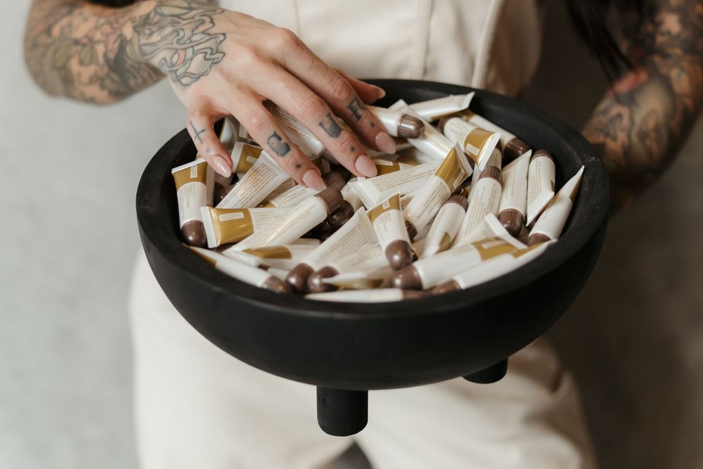 A tattooed hand reaches over an ashtray filled with discarded cigarette butts on a white surface.