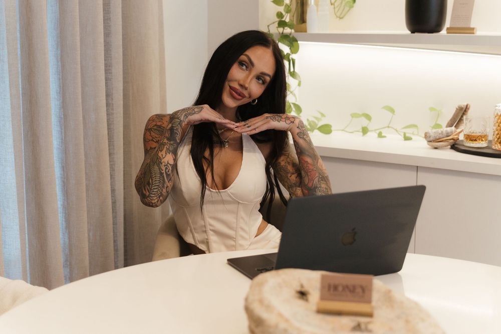 A person with tattoos works on a laptop while sitting at a white table in a bright modern home office setting.