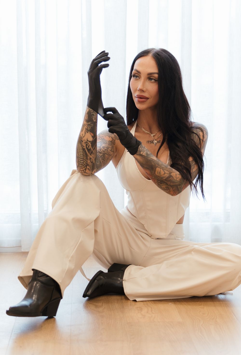 Model wearing cream-colored jumpsuit and black gloves sits on hardwood floor showcasing tattoo sleeves and dark boots.