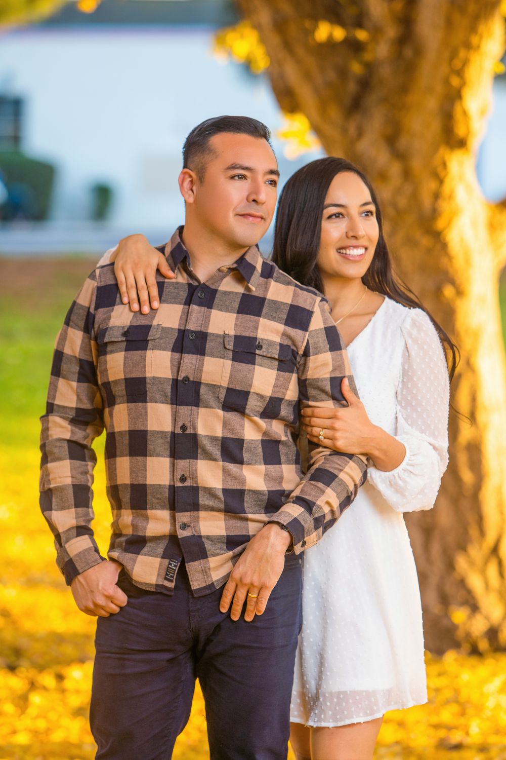 Couple in casual attire embracing under a golden-lit tree during sunset with autumn leaves on the ground.