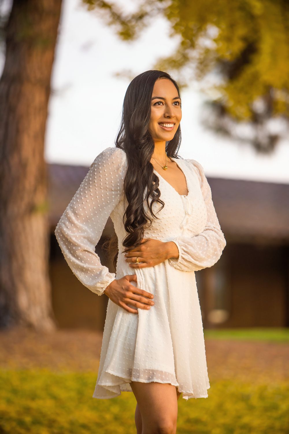 A person in a flowing white dress poses outdoors against a backdrop of autumn trees in warm golden light.