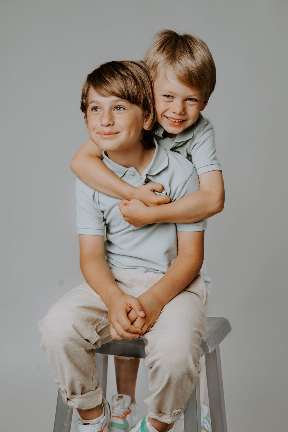 Two siblings in matching grey shirts share a playful hug while sitting on a stool.