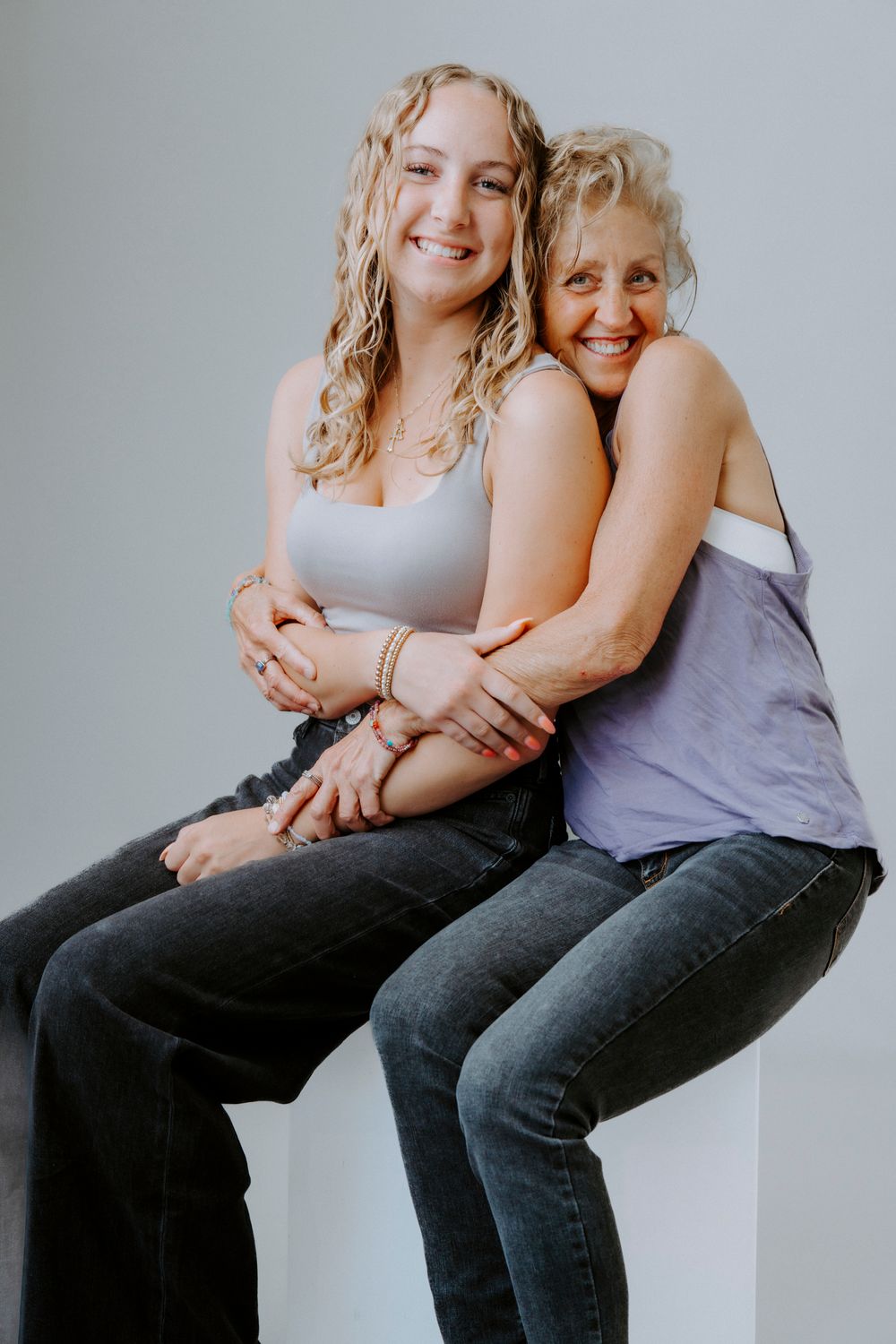 Two women sitting close together and embracing while wearing casual attire against a light background.