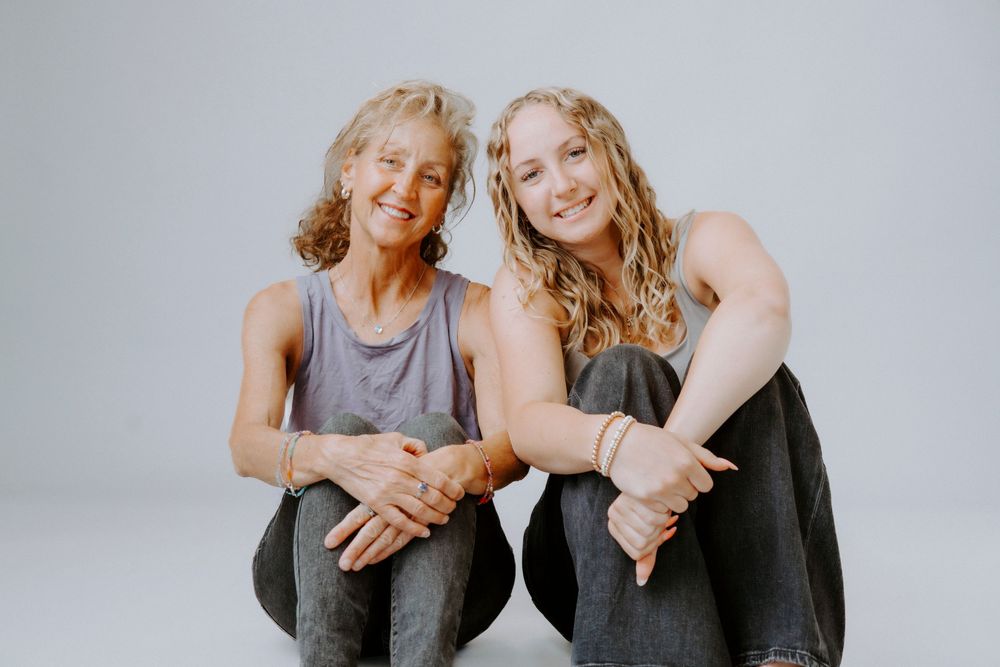 Two women in matching gray tops and jeans pose casually on a white backdrop.