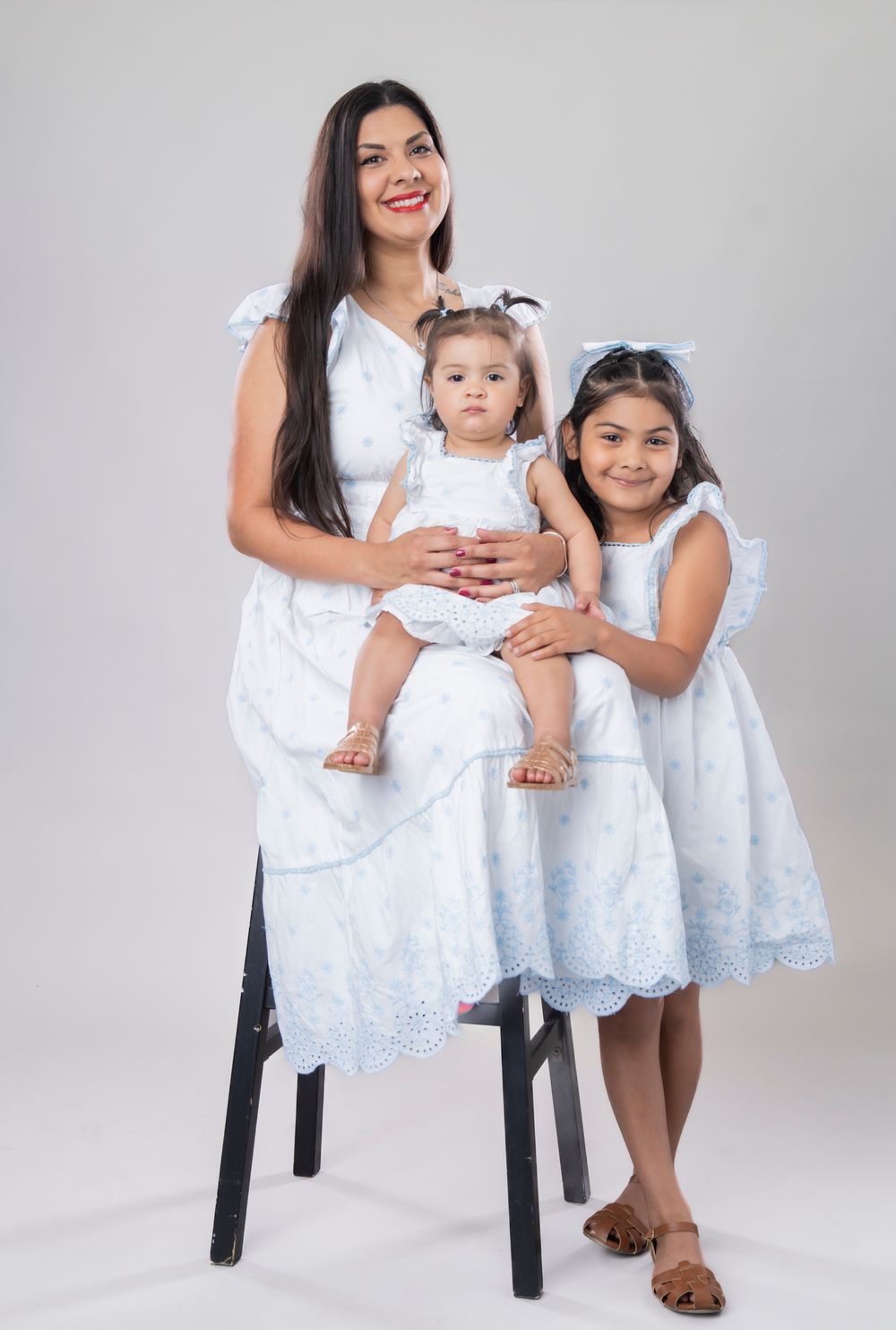 Three people in white lace dresses pose together on a stool against grey background.