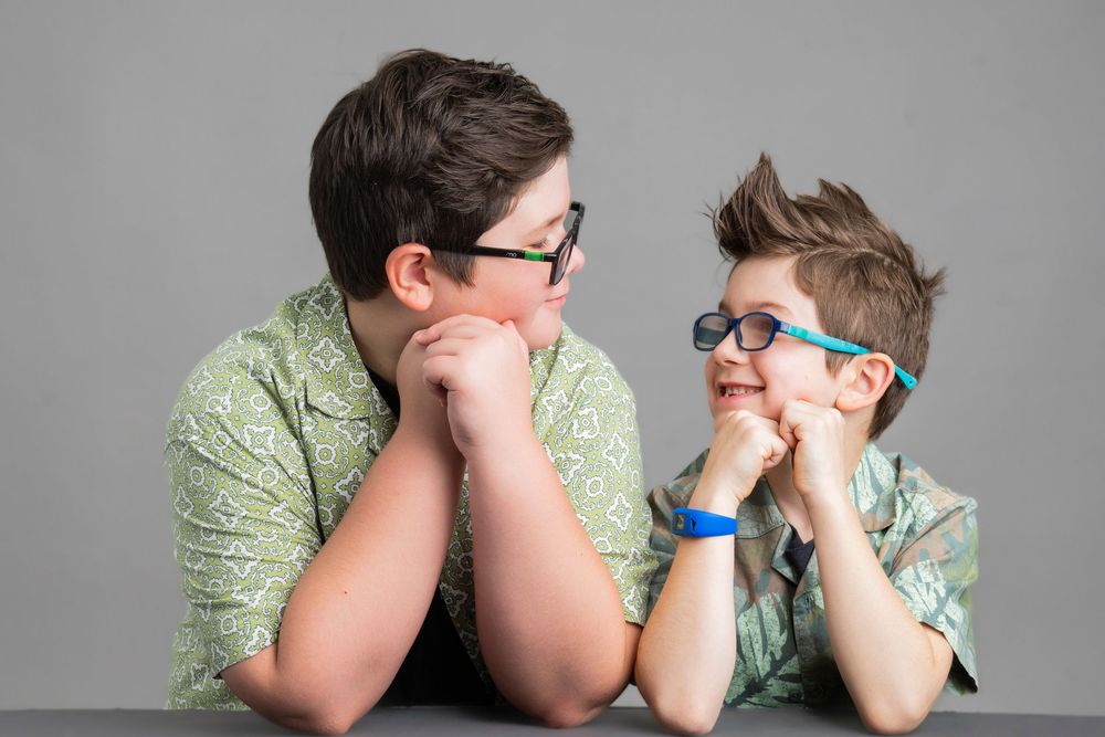 Two people in matching green shirts lean on their hands while sitting at a table wearing glasses.