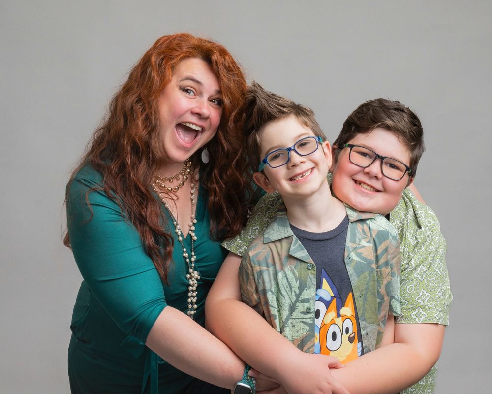 Portrait of three people posing together and laughing while wearing casual attire against a grey background.