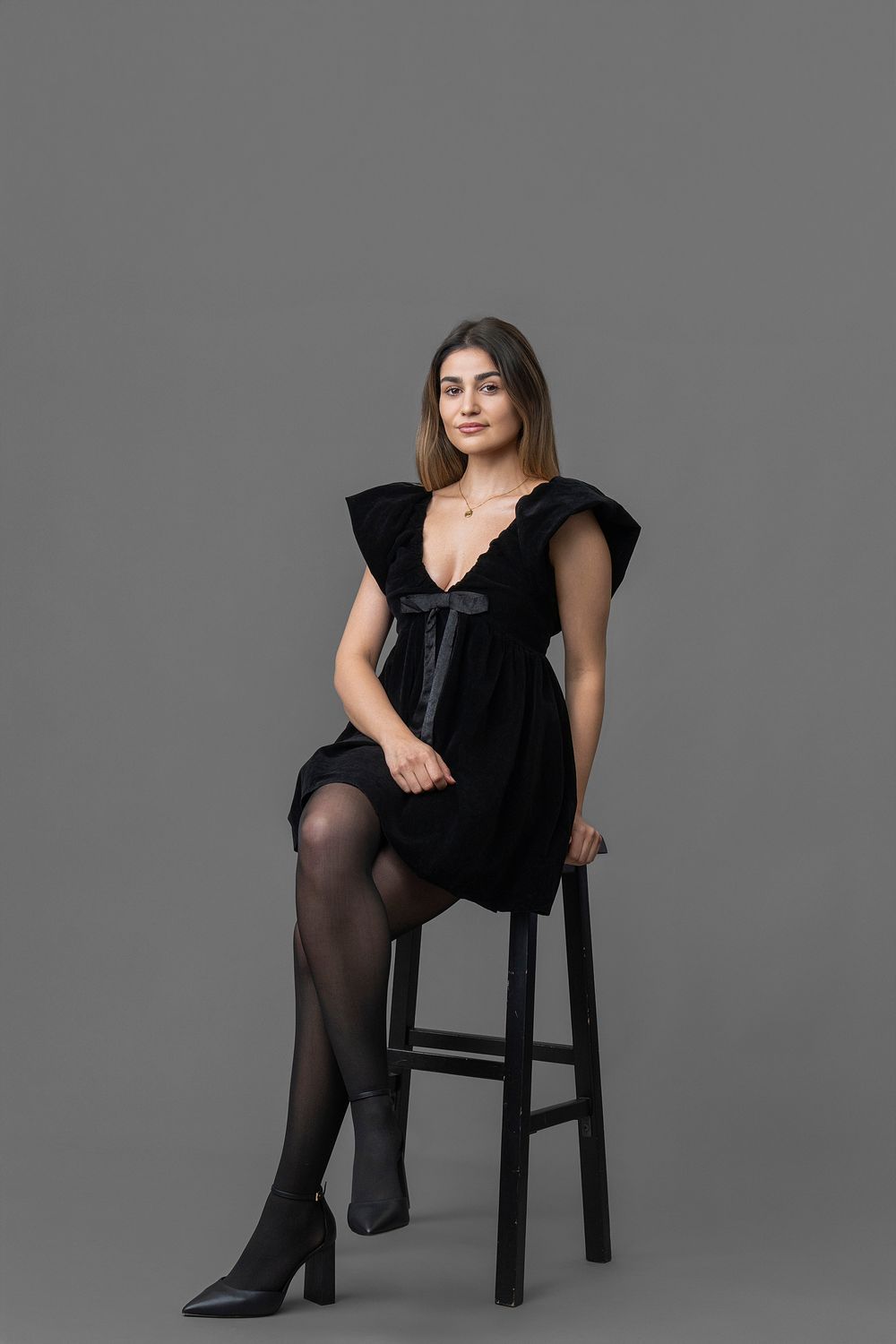 A figure in a black dress and tights poses elegantly on a wooden chair against a gray studio backdrop.