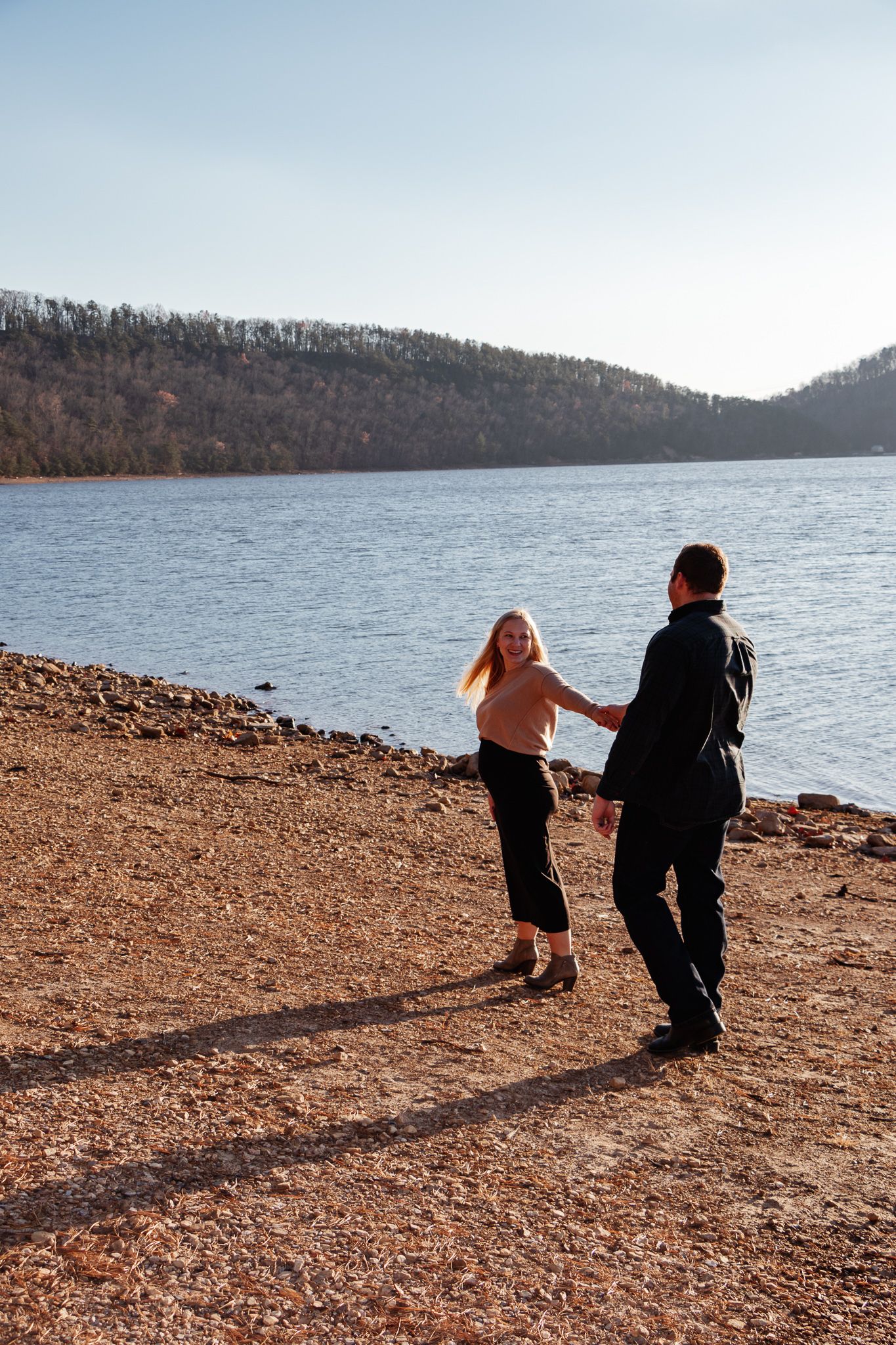 Couple holding hands and walking along a lakeshore, with mountains in the background and golden sunlight casting long shadows.