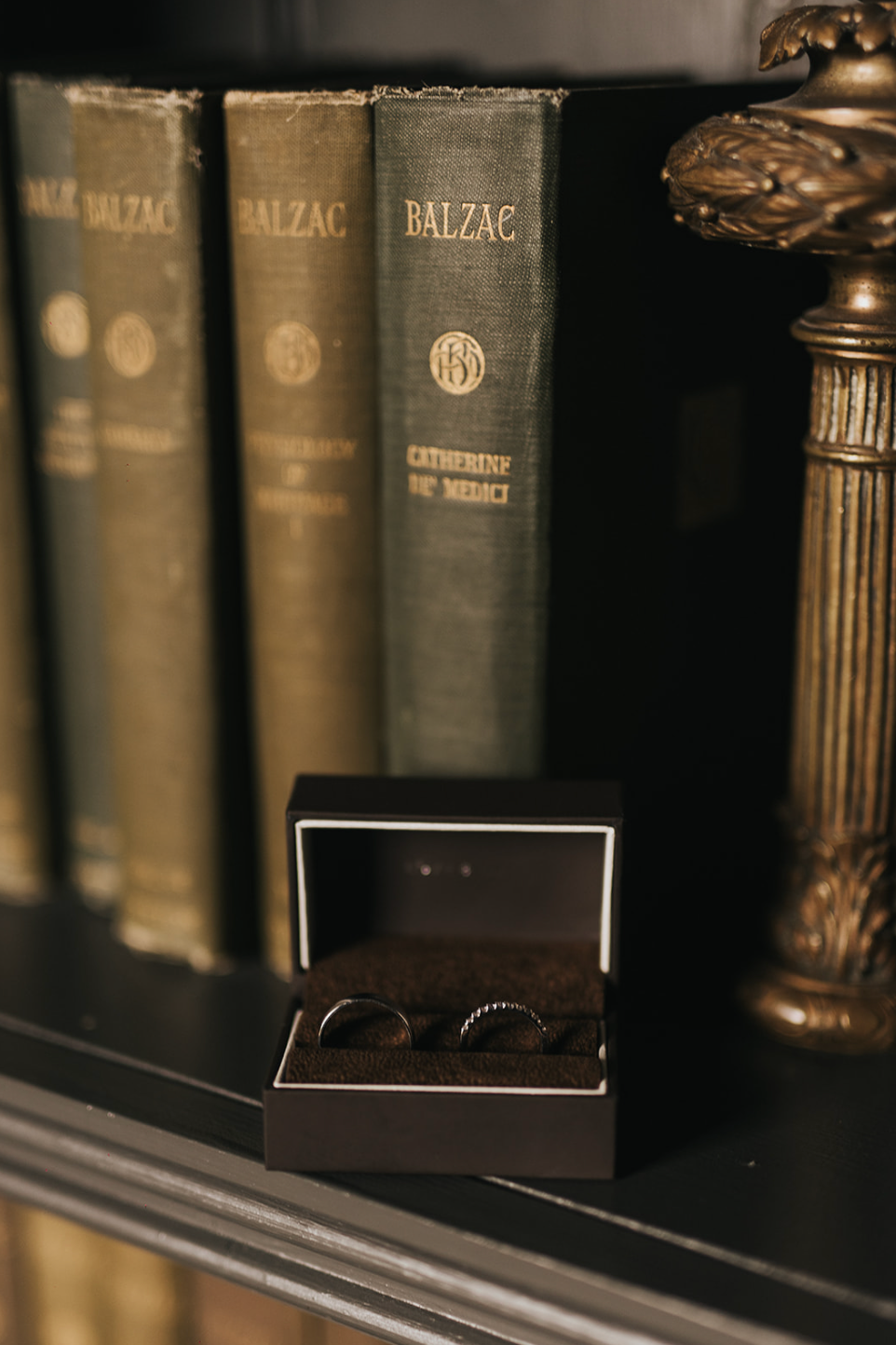 Wedding rings in a small box sit on a shelf with vintage Balzac books and decorative bookends.