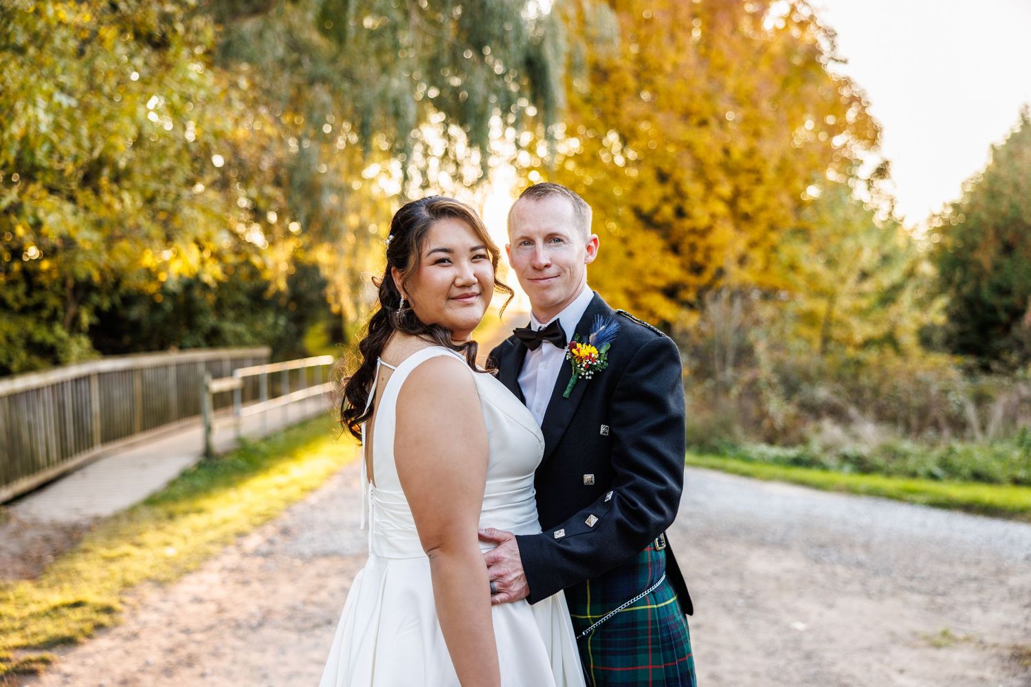 A joyful outdoor wedding photo featuring a couple on a path with autumn trees and golden sunlight in the background.