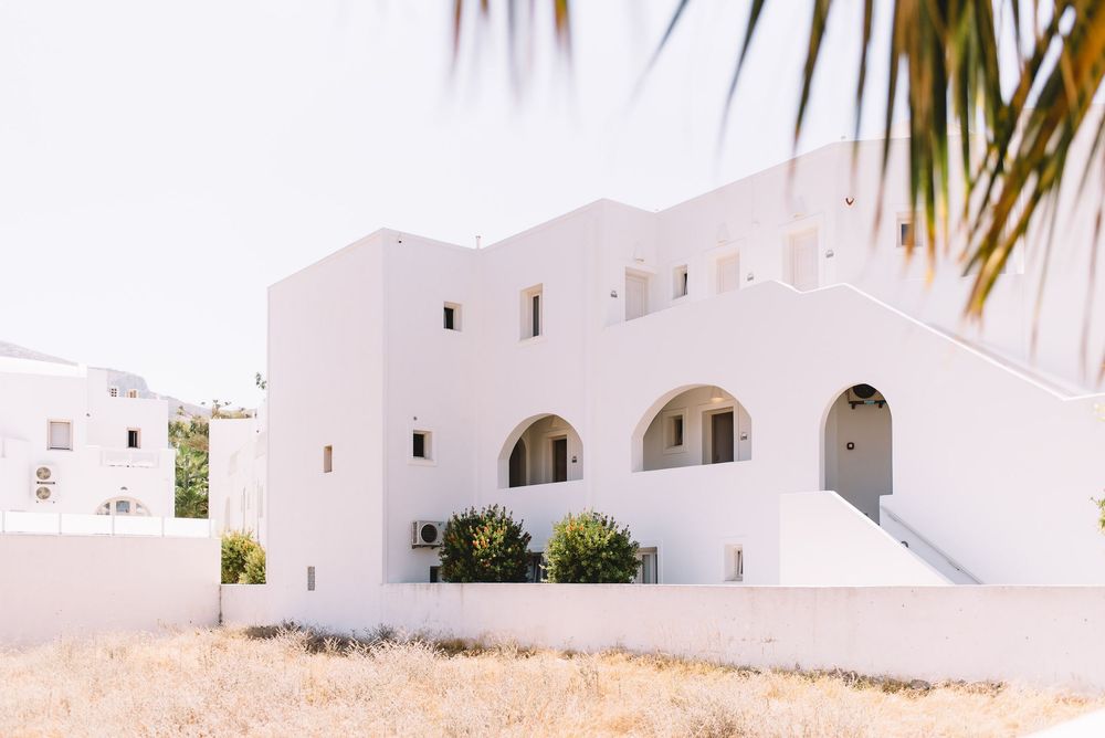 White Cycladic architecture building with arched doorways and lush green shrubs under bright sunlight in Greece.