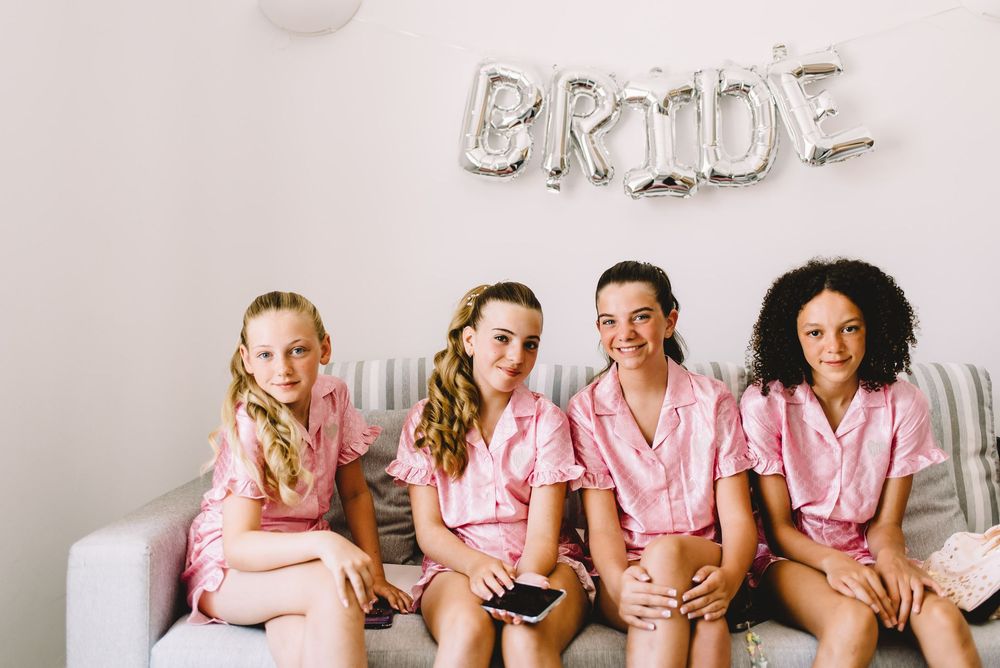 Four girls in matching pink pajamas sit on a couch beneath a silver BRIDE balloon banner at a bachelorette party.
