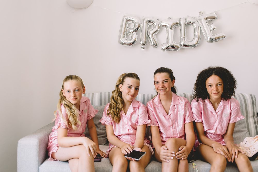 Bridesmaids wearing matching pink robes pose together under silver letter balloons spelling 'BRIDE'.