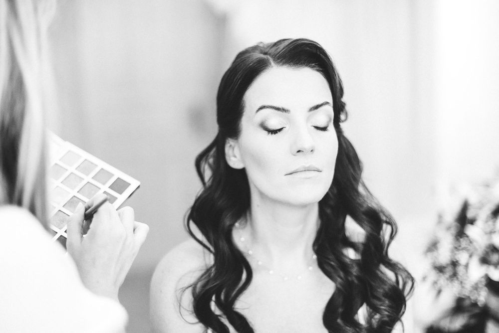 Black and white photo of a bride getting makeup applied by a professional artist before her wedding ceremony.
